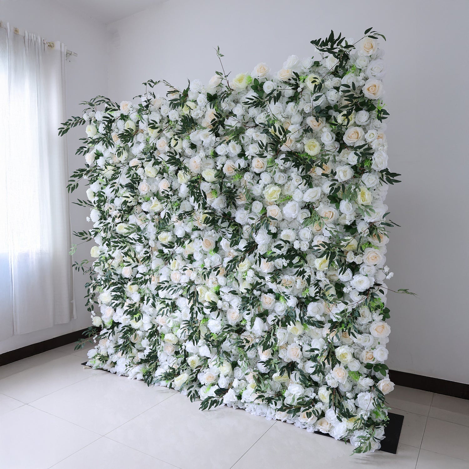 This image showcases a stunning floral wall by Valar Flowers. It features a dense arrangement of artificial flowers in soft shades of white and cream, interspersed with lush green foliage. The meticulously crafted blooms create a lush, elegant, and visually soothing display. Set indoors against a plain white wall and tiled floor, this floral wall is perfect for adding a touch of timeless beauty to events or spaces, exuding a refined and sophisticated atmosphere.