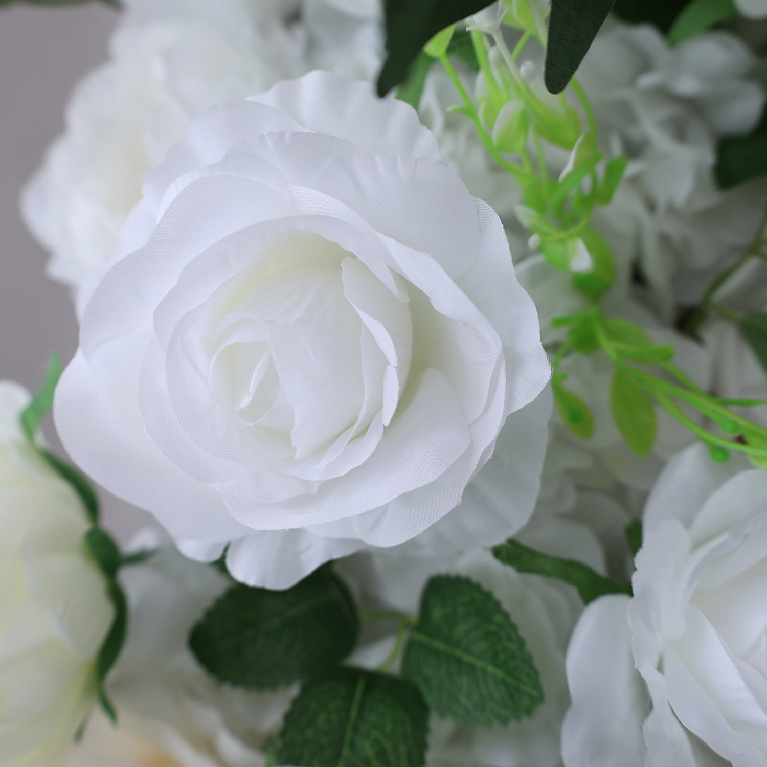 This image showcases a beautiful artificial white rose by Valar Flowers. The rose features delicate, layered petals with a soft, realistic texture. Surrounding it are other white blooms and lush green foliage, creating a cohesive and elegant floral arrangement. Perfect for adding a touch of timeless beauty to events or spaces, this piece exudes a refined and sophisticated charm, characteristic of Valar Flowers&