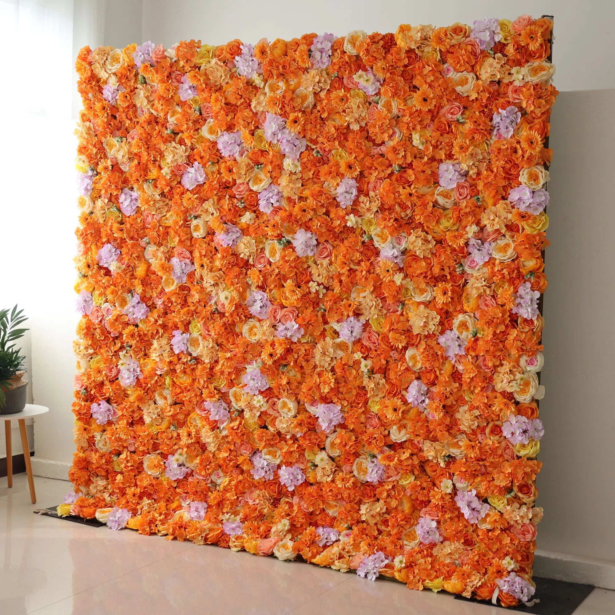 This image showcases a stunning floral wall by Valar Flowers, set indoors. Densely packed with artificial flowers in vibrant shades of orange, complemented by accents of purple, it creates a lively and eye - catching display. The flowers, including roses and other blooms, are arranged to form a lush, colorful panel. Placed on a light - colored tiled floor against a plain wall, this floral wall exudes elegance, making it perfect for decorative use in events or as a striking interior backdrop.