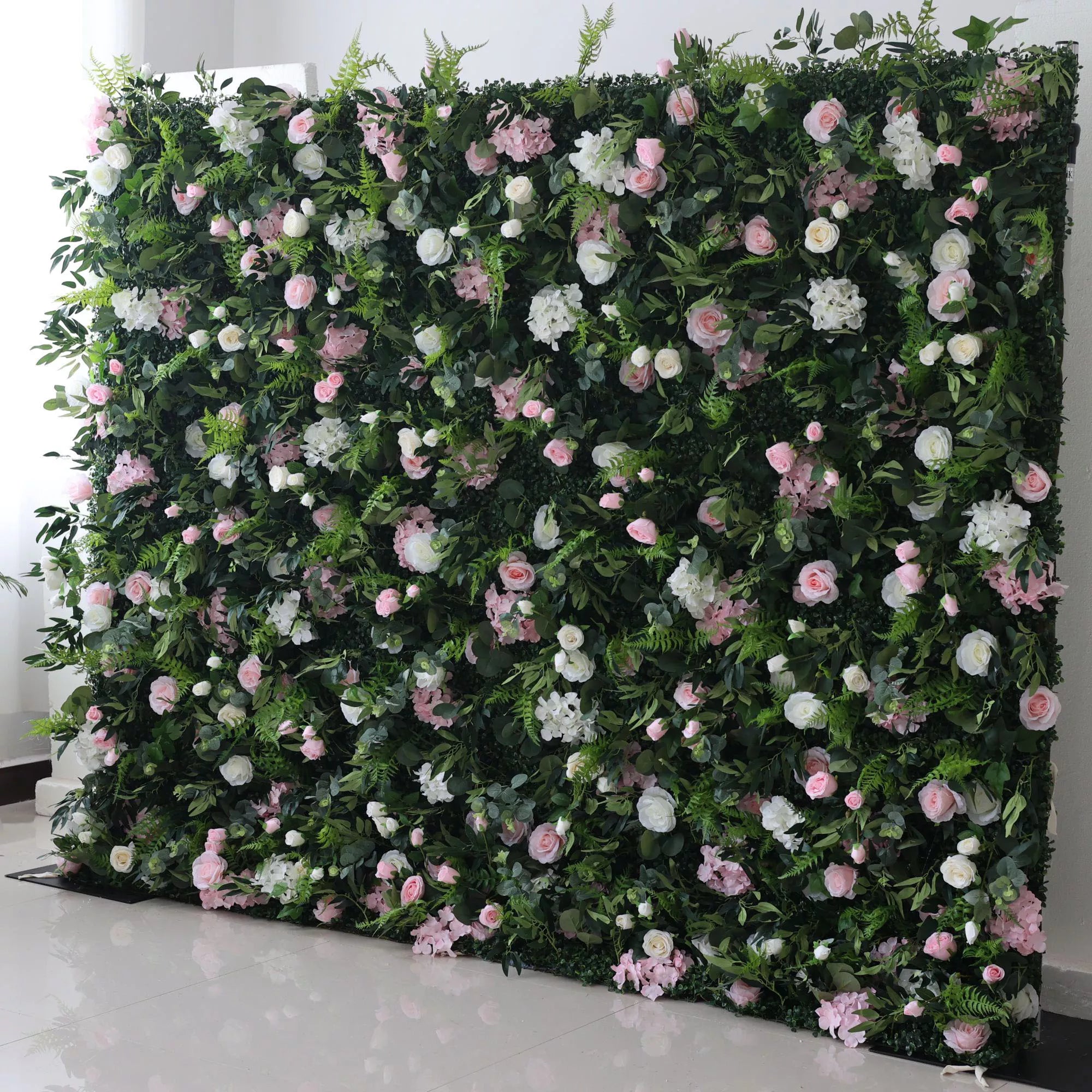 This lush floral wall features a dense, textured backdrop of deep green foliage, interspersed with a romantic mix of blush pink, soft white, and pale pink blooms—including roses, hydrangeas, and delicate filler flowers. The layered design creates a rich, three-dimensional texture, evoking a fresh, natural, and romantic atmosphere ideal for weddings, photo backdrops, or elegant celebrations.