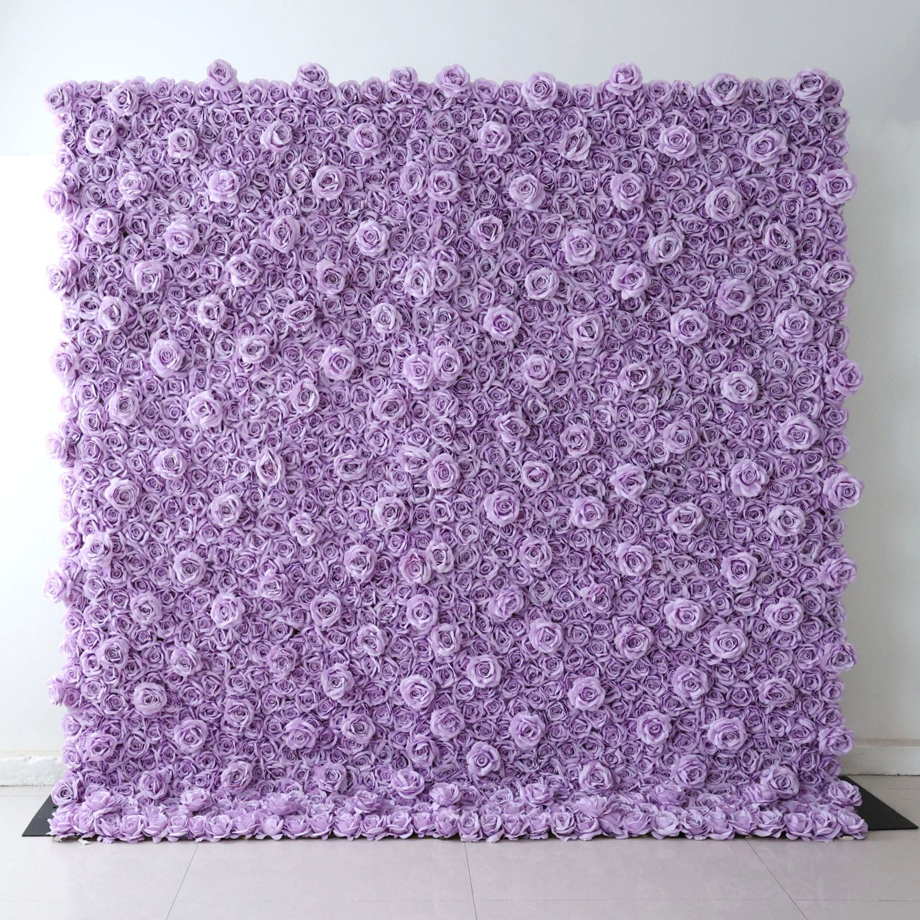 A square Valar Flowers floral wall panel, fully assembled against a plain white wall on light tile flooring, is entirely covered in dense, uniform lavender fabric roses. The seamless, textured design delivers a sleek, monochromatic aesthetic, perfect for minimalist weddings, baby showers, or purple-themed event photo backdrops.