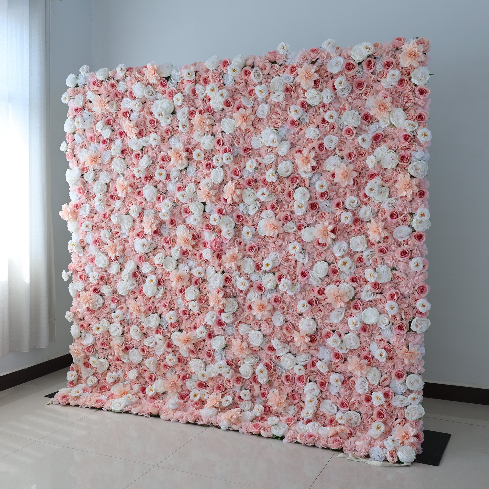 A large, square floral wall displays a vibrant mix of pink, blush, and white artificial flowers. Dense clusters of roses, peonies, and smaller blooms create a lush, romantic look. Set in a room with light - colored walls, a tiled floor, and a window with sheer curtains, it exudes elegance, perfect for weddings, photo backdrops, or upscale events needing a charming floral focal point.