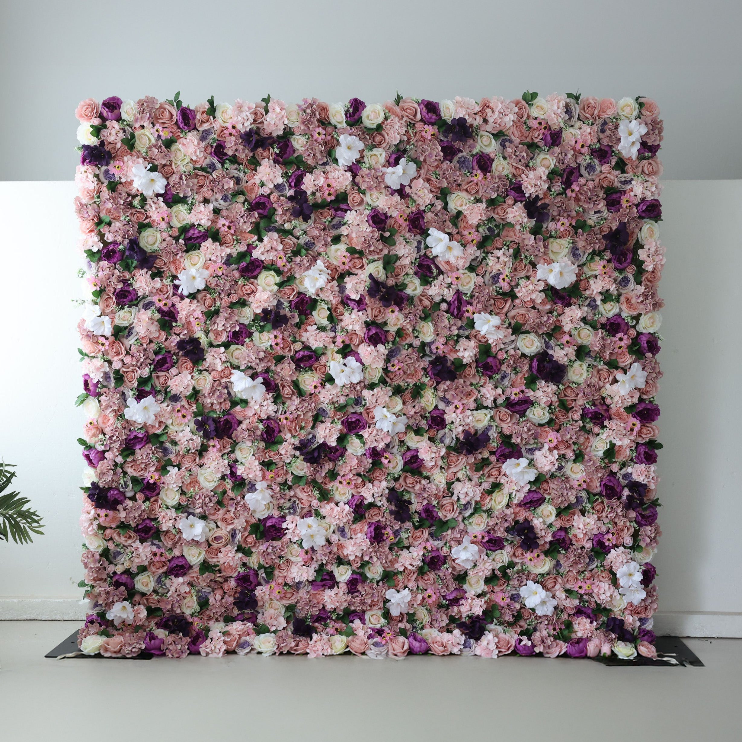 This opulent indoor floral backdrop features a dense, layered arrangement of artificial blooms in a romantic palette of blush, dusty rose, deep plum, and soft white. The rich mix of roses and filler flowers creates a lush, velvety texture that exudes vintage elegance and warmth. Set against a minimalist white wall and light floor, it is a stunning focal point for weddings, photoshoots, or upscale events.
