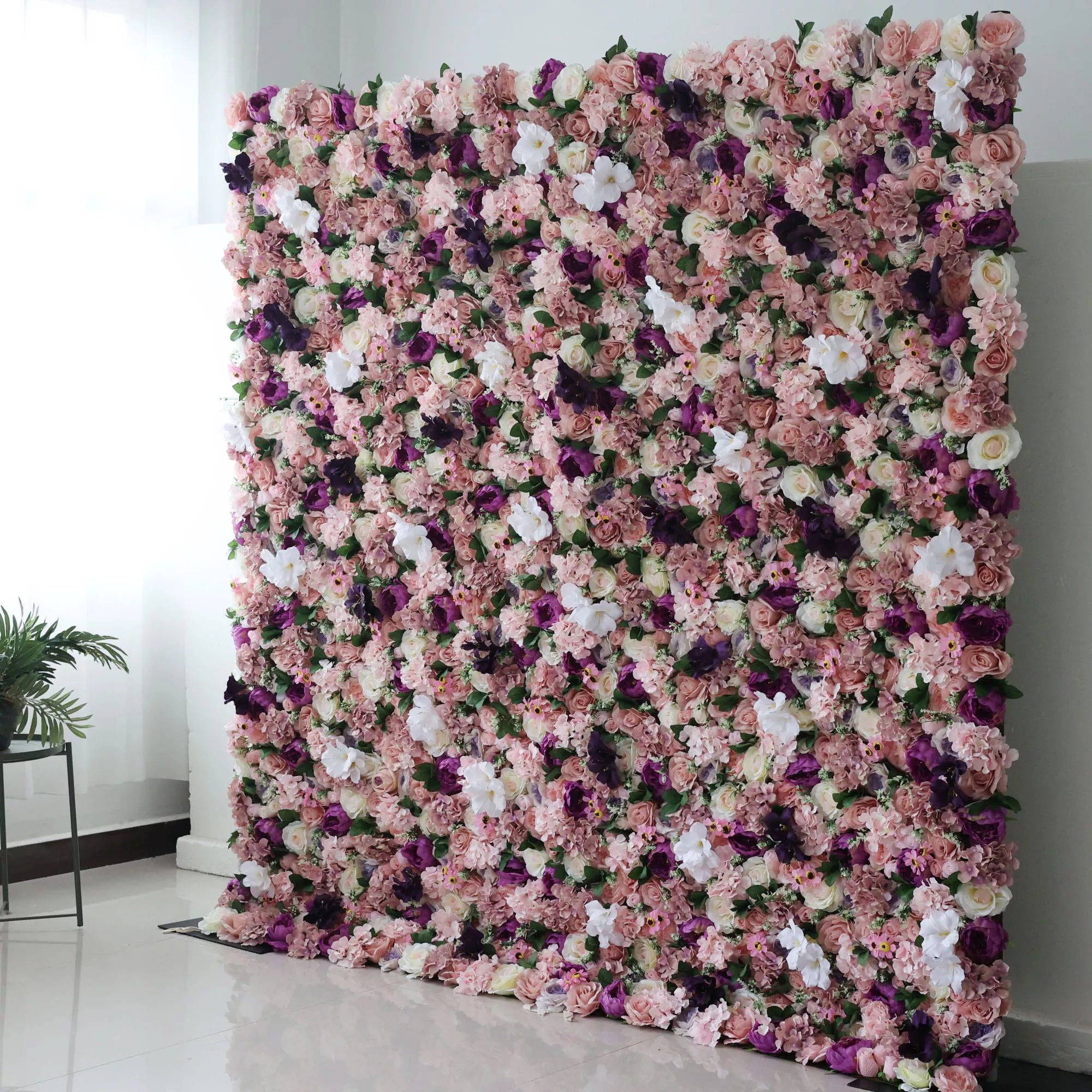 This opulent indoor floral backdrop features a dense, layered arrangement of artificial blooms in a romantic, vintage-inspired palette of blush, dusty rose, deep plum, and soft white. The rich mix of roses and filler flowers creates a lush, velvety texture that exudes warmth and elegance. Set against a minimalist white wall and light tiled floor, it is a stunning focal point for weddings, photoshoots, or upscale events.