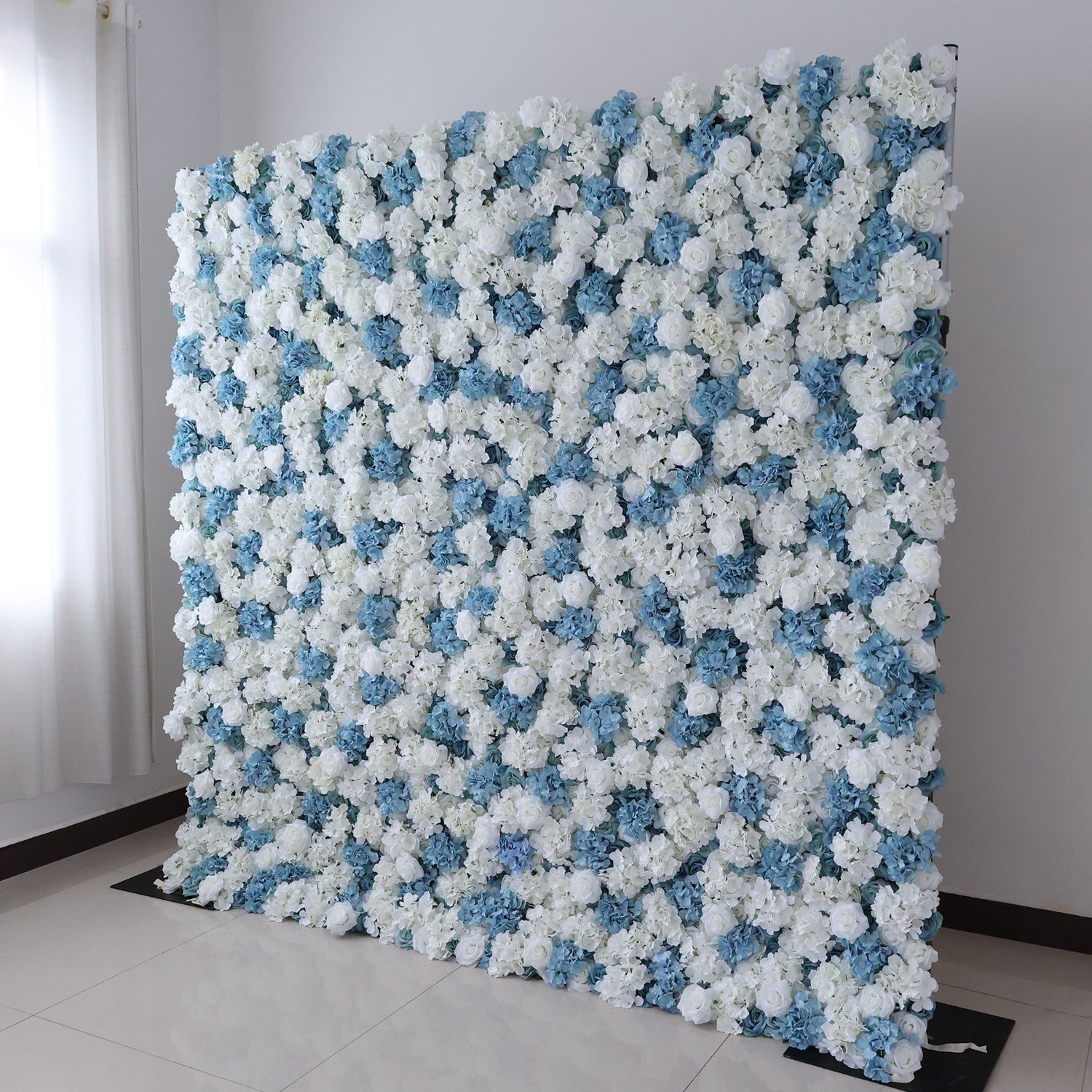 A large, square floral wall showcases a beautiful blend of white and blue artificial flowers. Dense clusters of blooms create a lush, elegant display. Set in a room with light - colored walls, a tiled floor, and a window with sheer curtains letting in soft light, it exudes a fresh and sophisticated vibe, perfect for events like weddings, parties, or as a decorative photo backdrop.