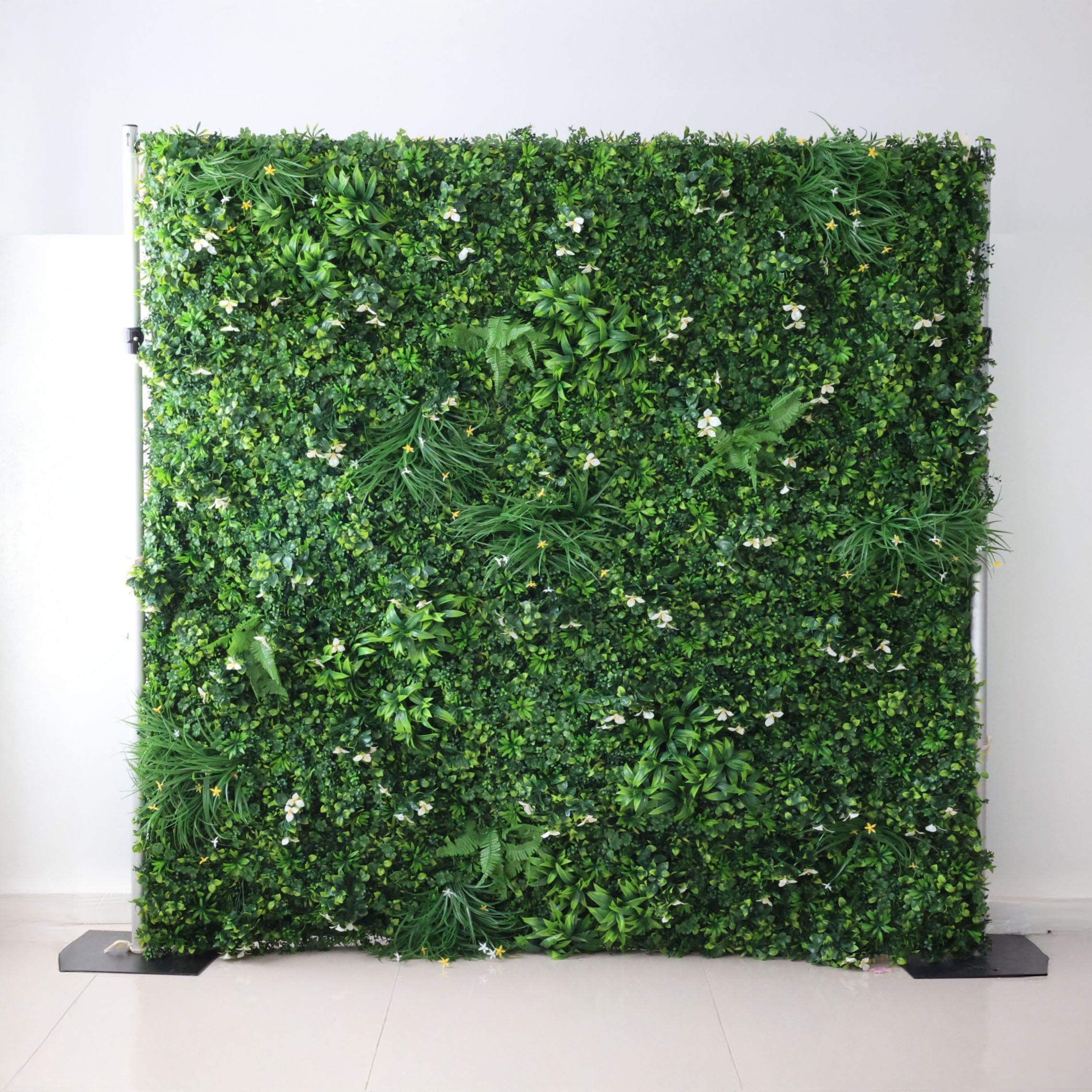 This image showcases a lush artificial greenery wall by ValarFlowers, set indoors against a plain white wall. It features a dense arrangement of varied green foliage, interspersed with delicate white blooms and subtle yellow accents, along with ferns and textured plants. The realistic textures and diverse elements create a rich, natural, and visually captivating display, perfect for adding organic elegance to interiors or events, exuding a serene and sophisticated atmosphere.