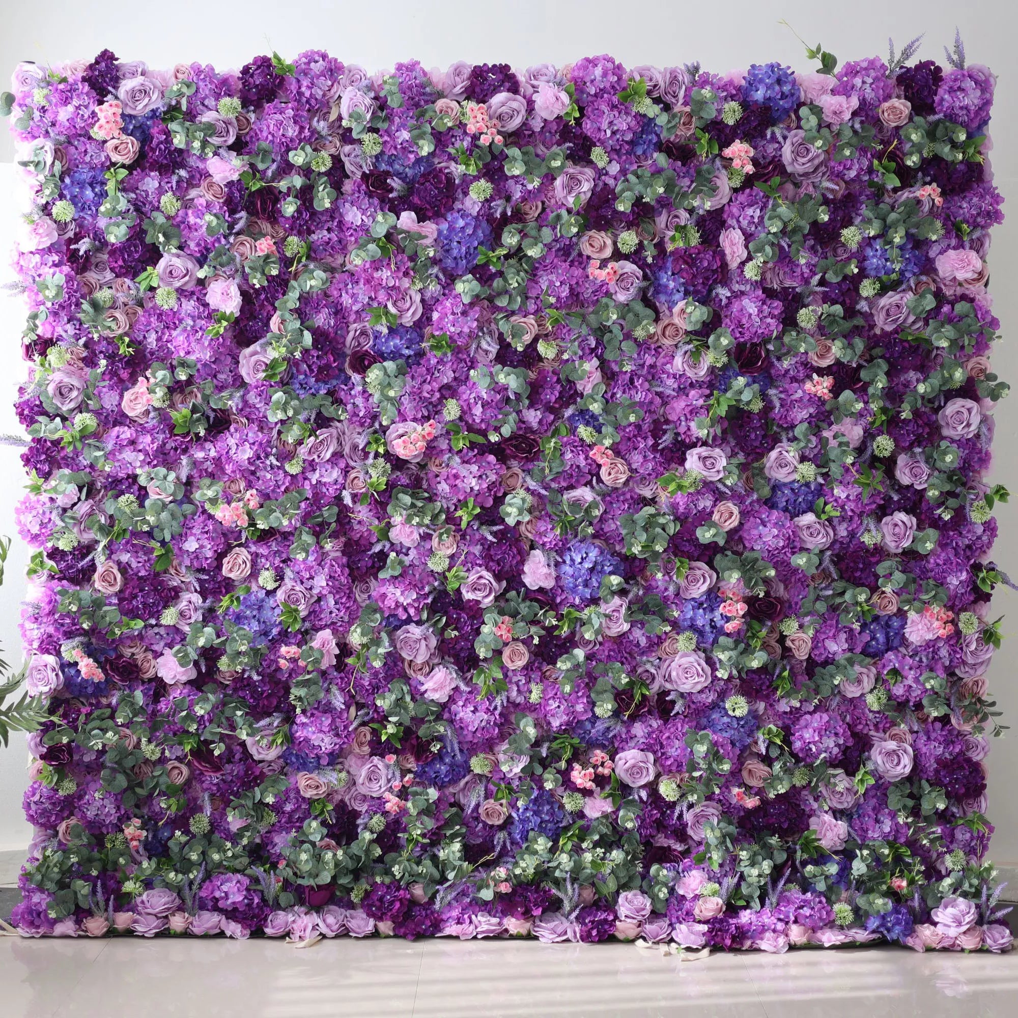 This image features a large, lush floral wall dominated by various shades of purple, from deep violet to soft lavender, along with hints of blue and pink blooms. Interspersed with green foliage, the dense arrangement creates a rich, vibrant, and elegant display. Set against a plain indoor background with a shiny floor, the floral wall exudes a sense of luxury and natural beauty, perfect for decorative purposes in events or as a striking backdrop.