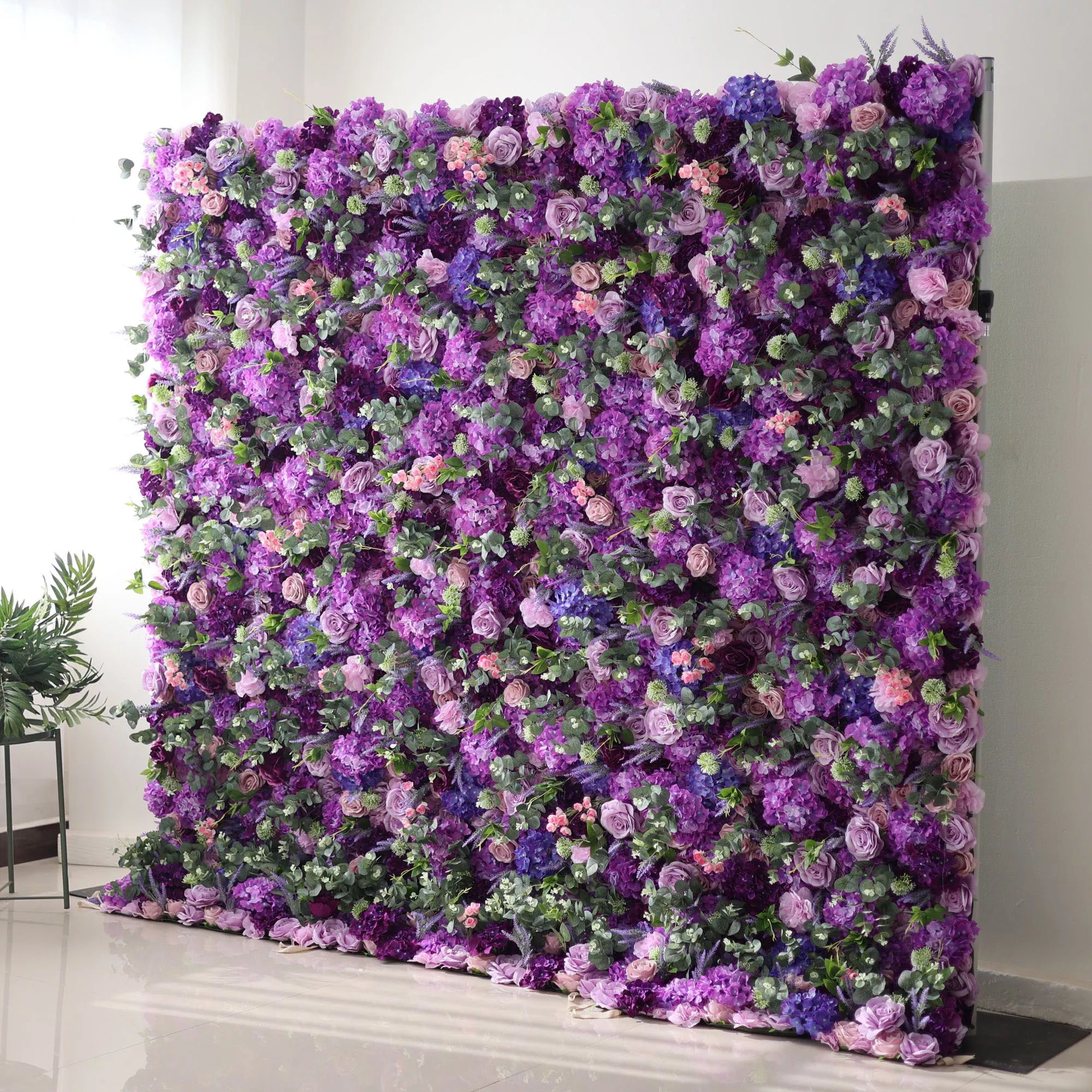 This image features a large, lush floral wall dominated by various shades of purple, from deep violet to soft lavender, along with hints of blue and pink blooms. Interspersed with green foliage, the dense arrangement creates a rich, vibrant, and elegant display. Set against a plain indoor background with a shiny floor, the floral wall exudes a sense of luxury and natural beauty, perfect for decorative purposes in events or as a striking backdrop.