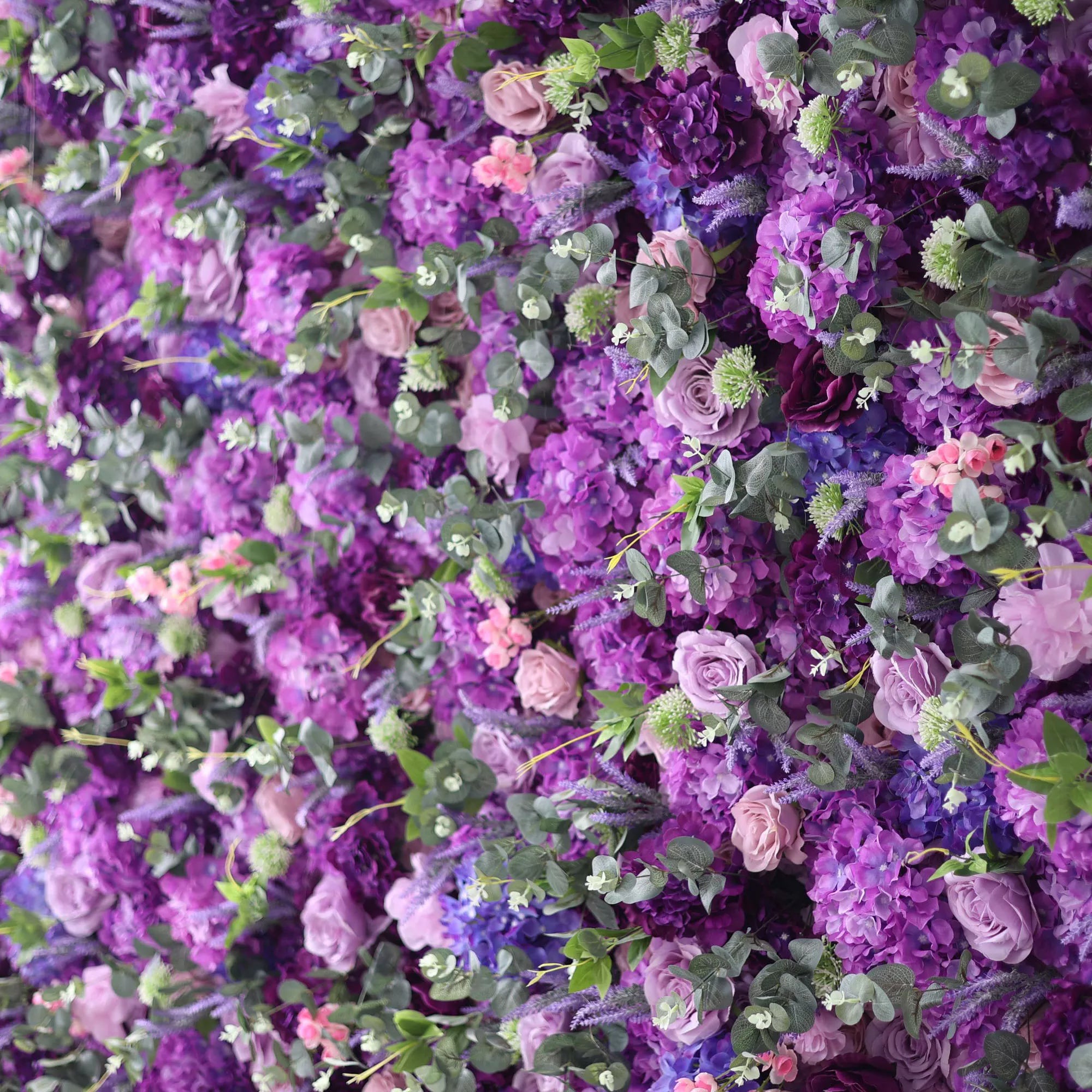 This image showcases a lush floral wall brimming with blooms in various shades of purple, from deep violet to soft lavender, along with hints of pink and blue flowers. Interspersed with green foliage, the dense arrangement creates a rich, vibrant, and elegant display. The intricate mix of petals and leaves gives a sense of natural abundance and beauty, making it a perfect decorative element for events or as a stunning backdrop.