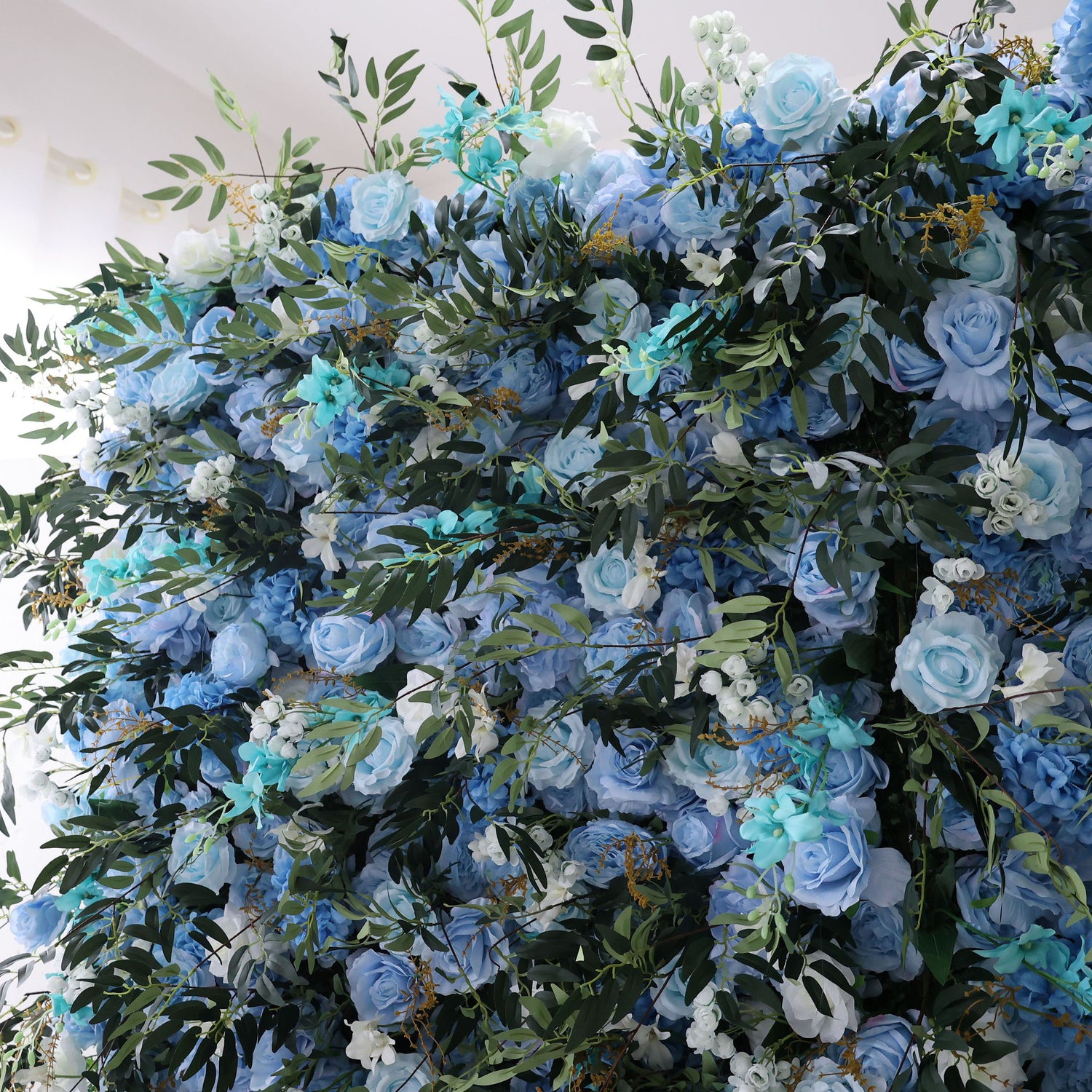 This image showcases a stunning floral wall by Valar Flowers. It features a dense arrangement of artificial flowers in various shades of blue, from light sky blue to deeper hues, complemented by white blooms. Lush green foliage is interspersed, creating a lush, serene, and visually captivating display. Perfect for adding an elegant, nature - inspired touch to events or interior spaces, it exudes a sense of tranquility and beauty.