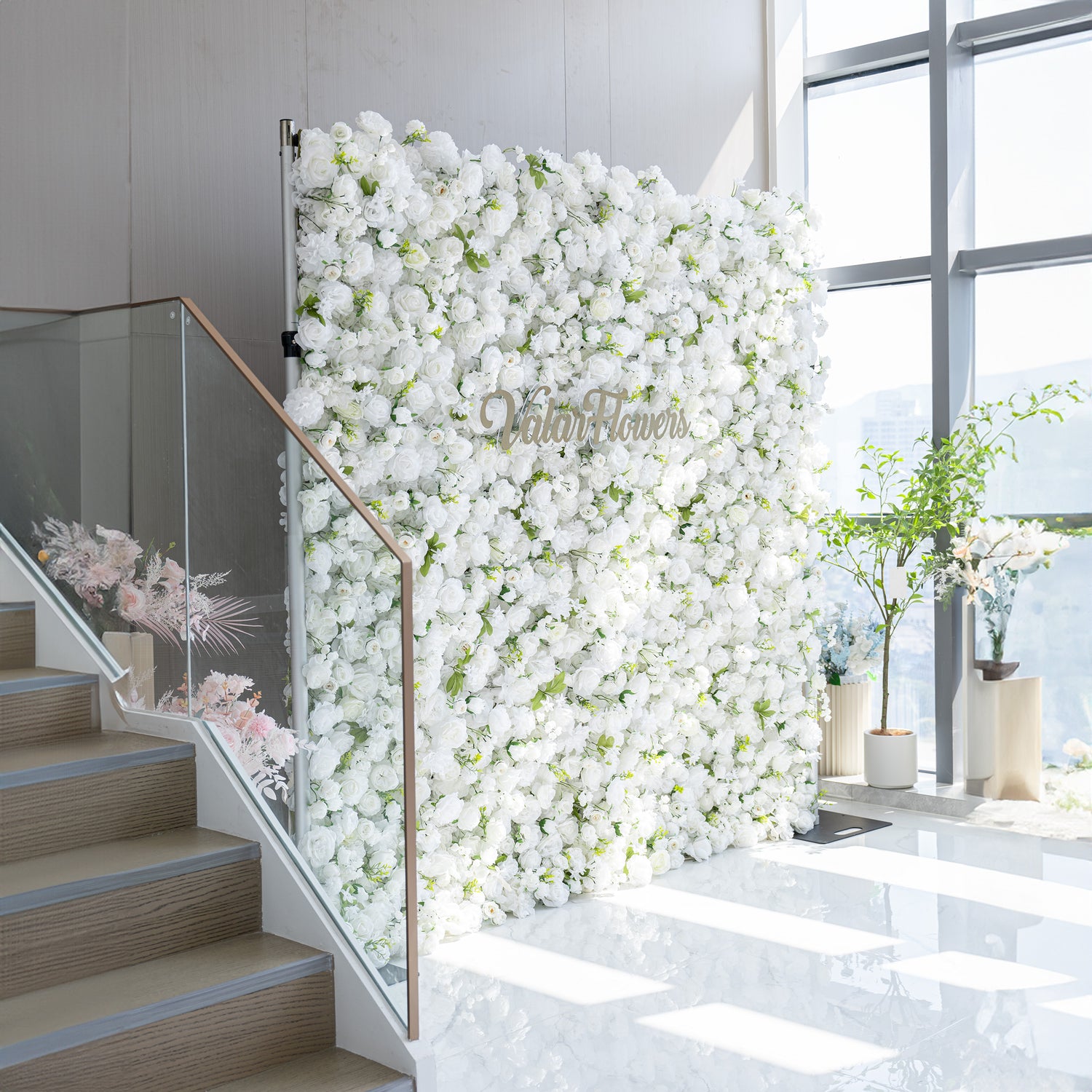This floral backdrop is densely covered with artificial white flowers, mainly roses and some other assorted blooms, interspersed with green leaves. The all - white color palette, complemented by the greenery, creates a clean, elegant, and sophisticated style. The golden “ValarFlowers” sign adds a touch of luxury, making it perfect for formal events.