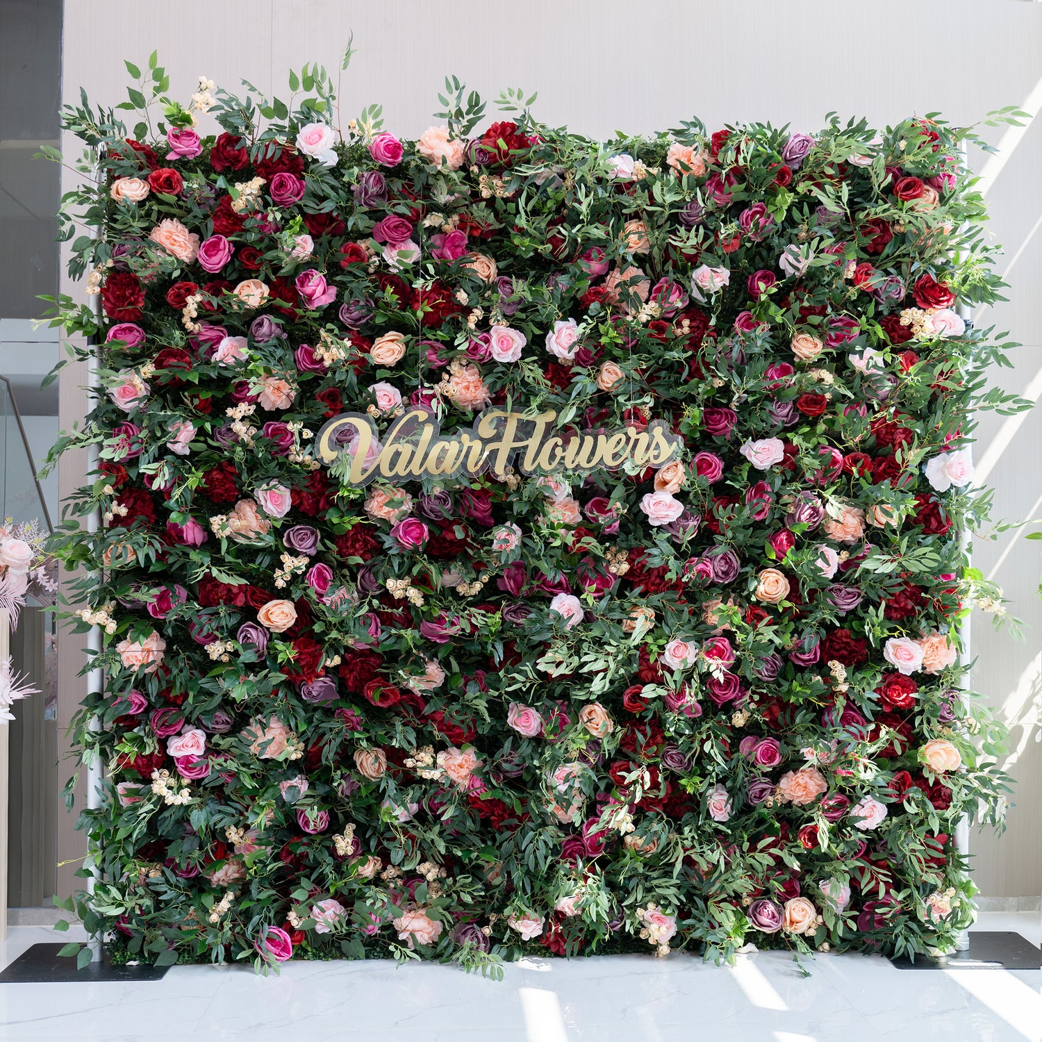 A large, lush artificial flower wall showcases a vibrant mix of roses in pink, red, purple, and peach, interspersed with green foliage and small white flowers. At its center, a gold “ValarFlowers” sign adds a branding touch, creating an elegant, eye - catching decorative backdrop.