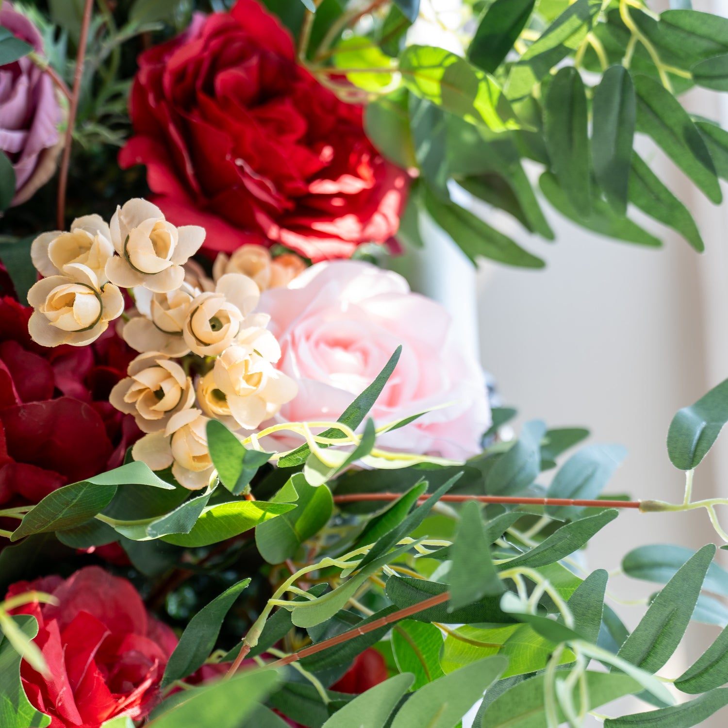 A close - up of artificial flowers: deep red roses, soft pink blooms, and tiny cream - colored buds, all surrounded by green foliage. The delicate petals and lush leaves create a romantic, textured arrangement, perfect for decorative displays.
