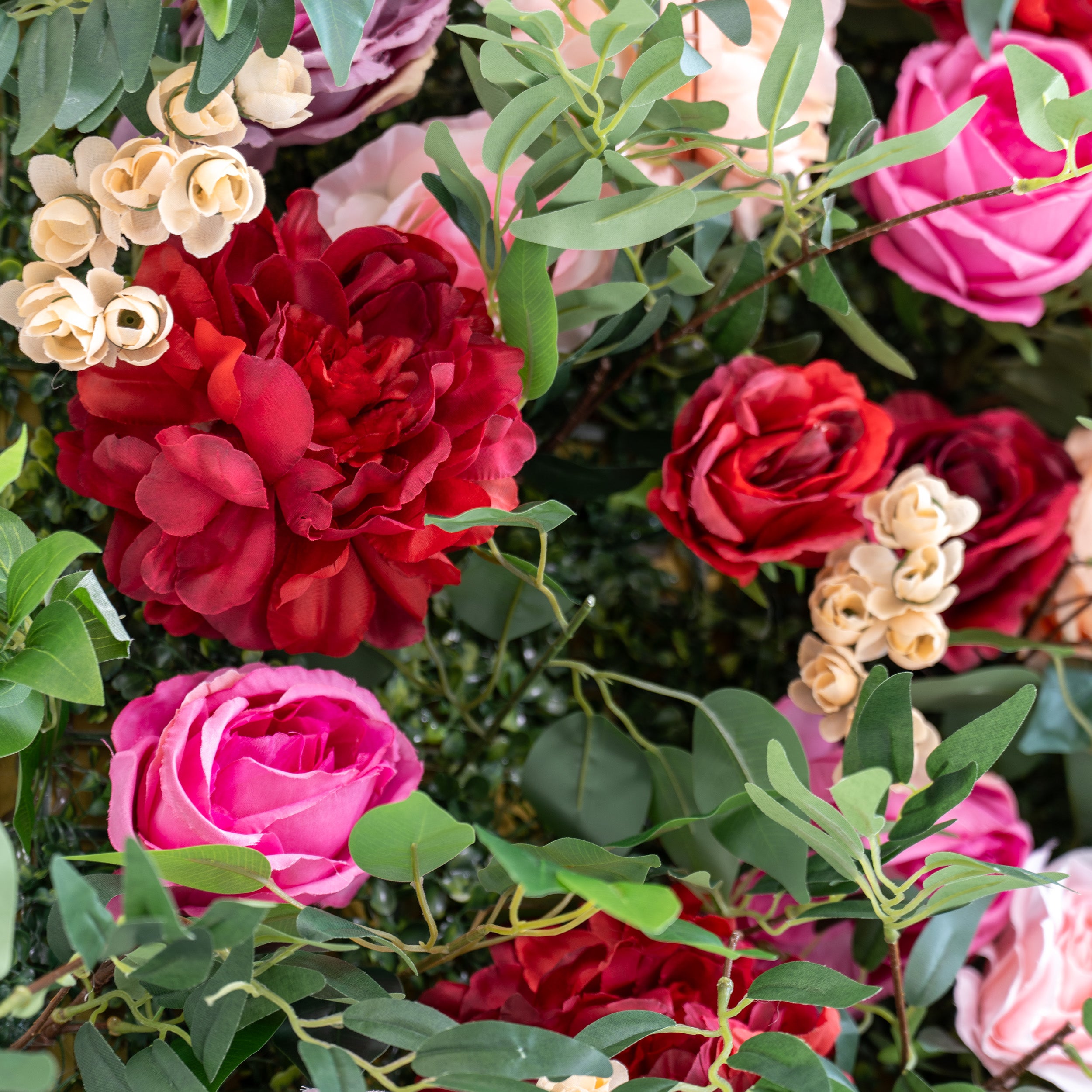 A close - up of artificial flowers features vibrant red peonies, pink roses, and tiny cream - colored buds, all surrounded by green foliage. The lush, layered petals and varied hues create a romantic, decorative arrangement, perfect for backdrops or floral displays.