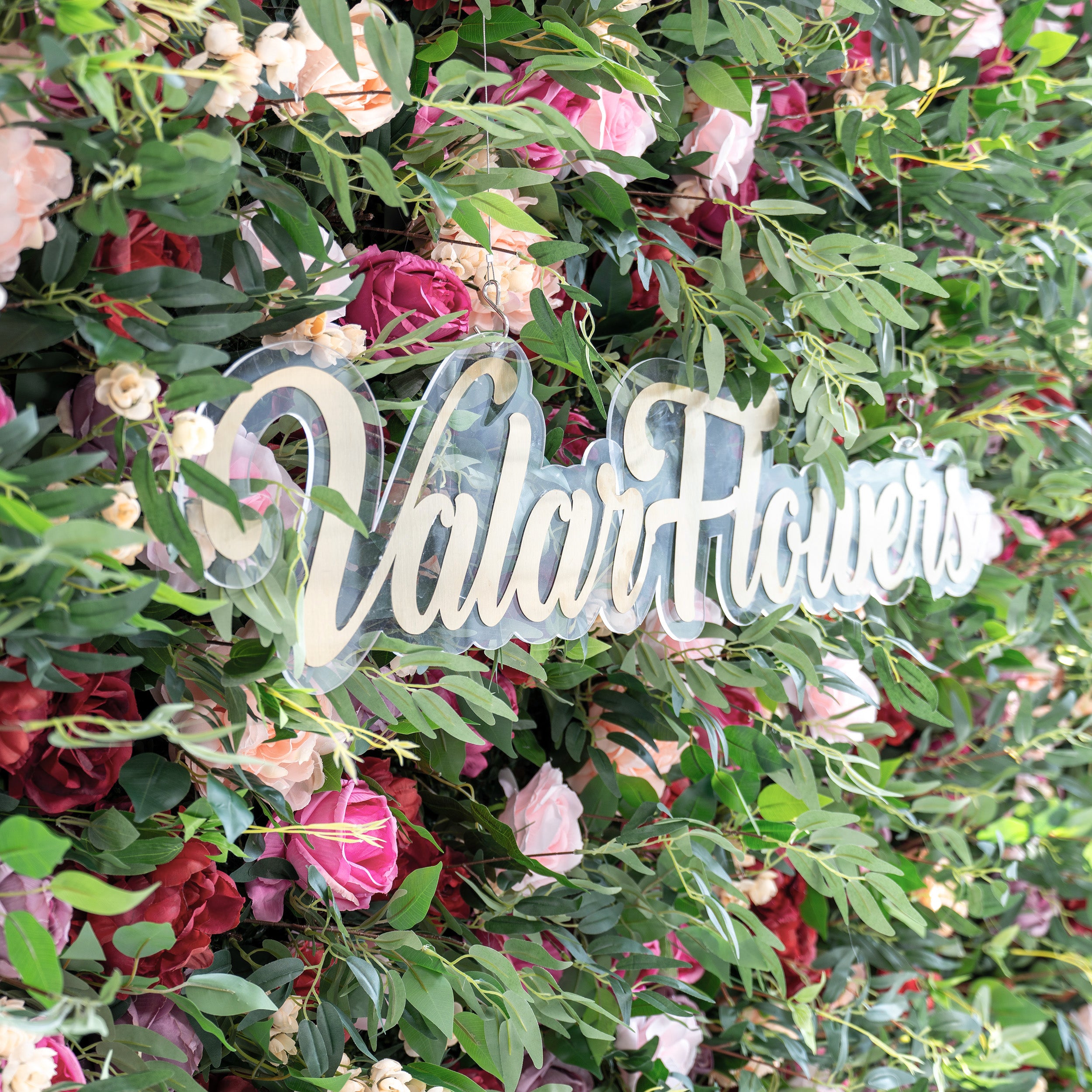 A close - up of a clear acrylic sign with gold “ValarFlowers” script, nestled in a lush artificial flower wall. The wall features pink, red, and peach roses, plus green foliage, creating a vibrant, decorative backdrop that highlights the branding element.