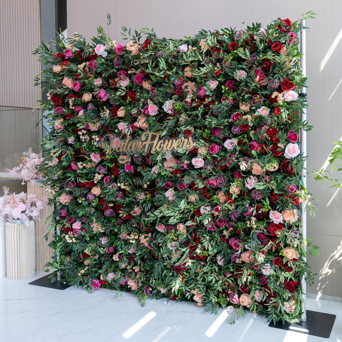 A large, lush artificial flower wall features a vibrant mix of roses in pink, red, purple, and peach, plus green foliage and small white flowers. A gold “ValarFlowers” sign stands out in the center. It’s an eye - catching, elegant backdrop, perfect for events or decorative displays.