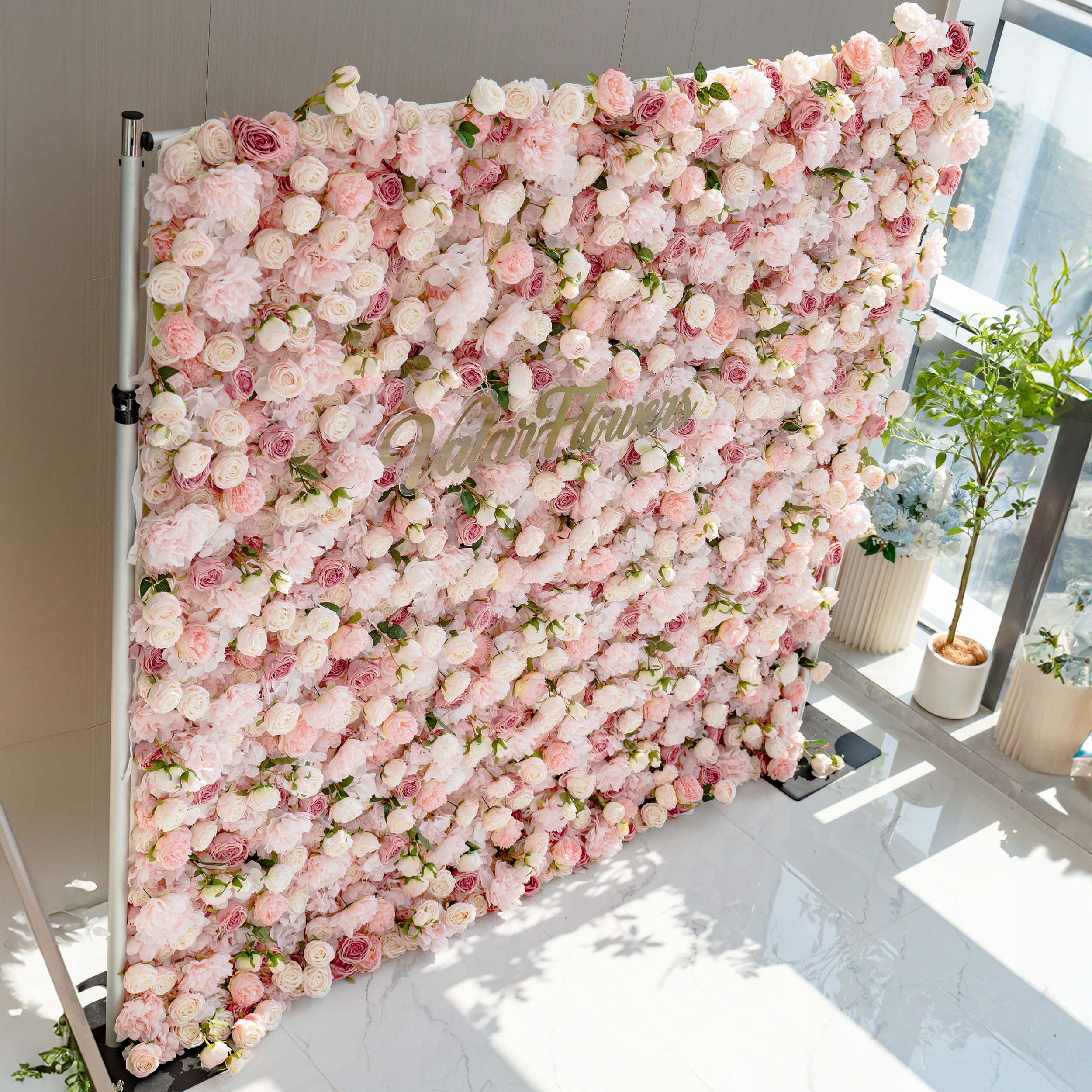 A stunning flower wall filled with a variety of pink and white roses. The wall is labeled "ValarFlowers" and is set against a modern interior with potted plants nearby.