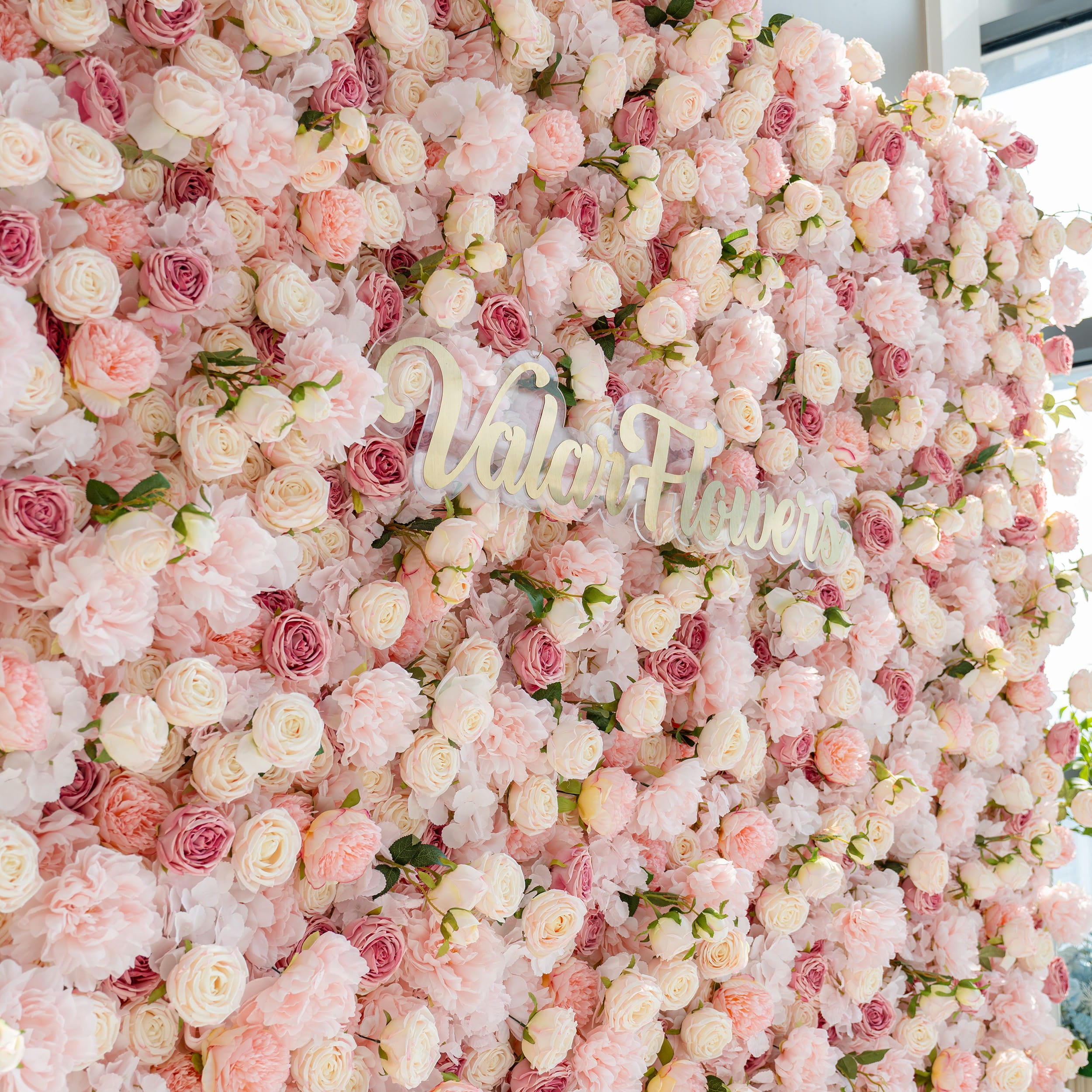 A beautiful flower wall composed of various pink and white roses and peonies. The words "Valar Flowers" are prominently displayed in the center, creating a romantic and elegant atmosphere.