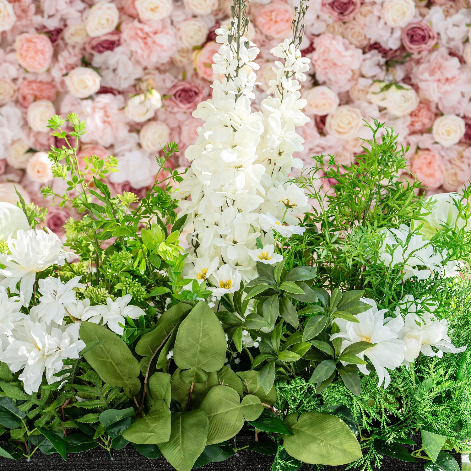 The floral arrangement features a variety of white flowers, including lilies and orchids, surrounded by lush green foliage. It is set against a backdrop of pink and cream roses. This setup is perfect for weddings, creating a romantic and elegant atmosphere.