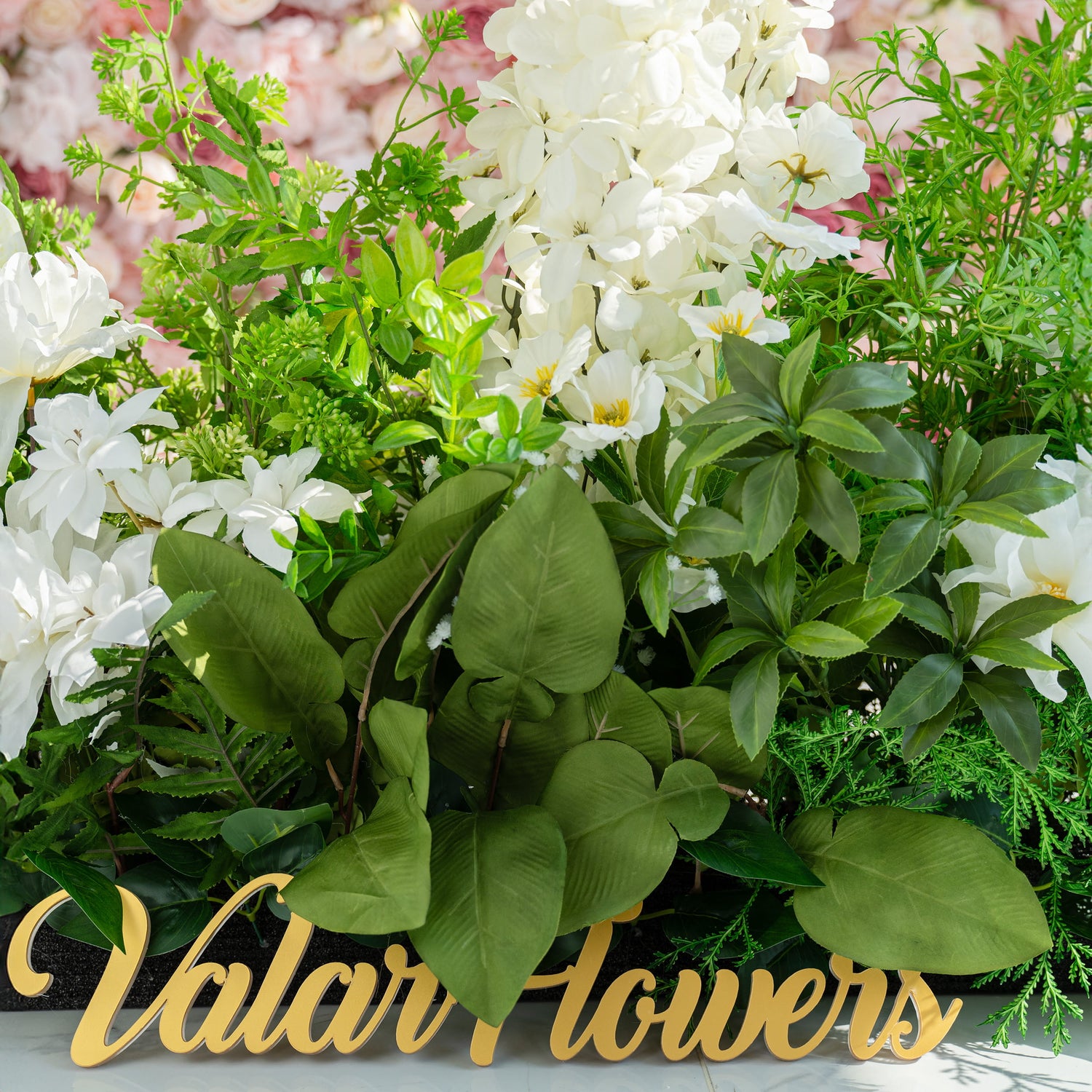 The floral arrangement features a variety of white flowers, including lilies and orchids, surrounded by lush green foliage. It is ideal for weddings, creating a romantic and elegant atmosphere. The sign "Valar Flowers" adds a charming touch.