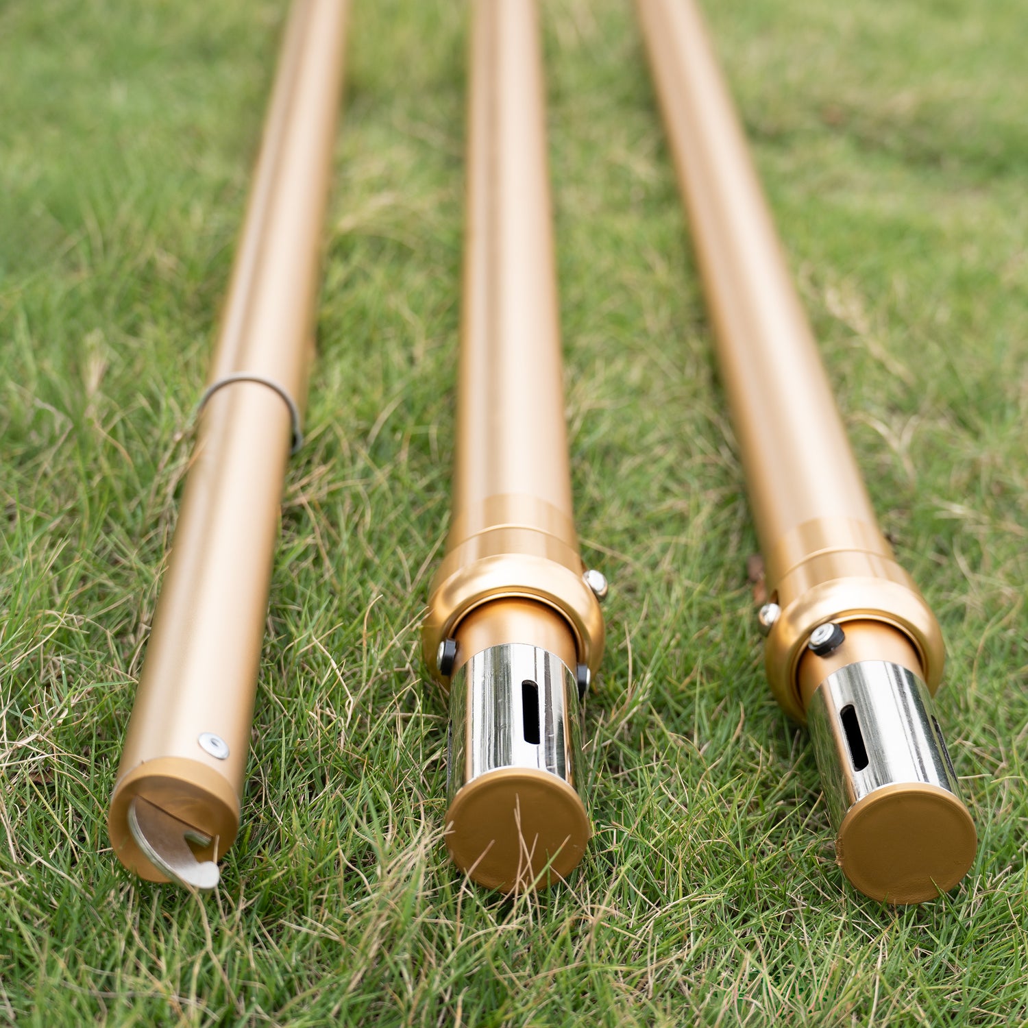This image shows three golden - colored metal poles lying on a grassy surface. The poles appear to be parts of a backdrop stand or similar structure. Two of the poles have metallic tops with vertical slits and are capped with gold - colored ends, while the third pole has a curved attachment at its end. The setup suggests they are ready for assembly, likely for event decoration purposes.