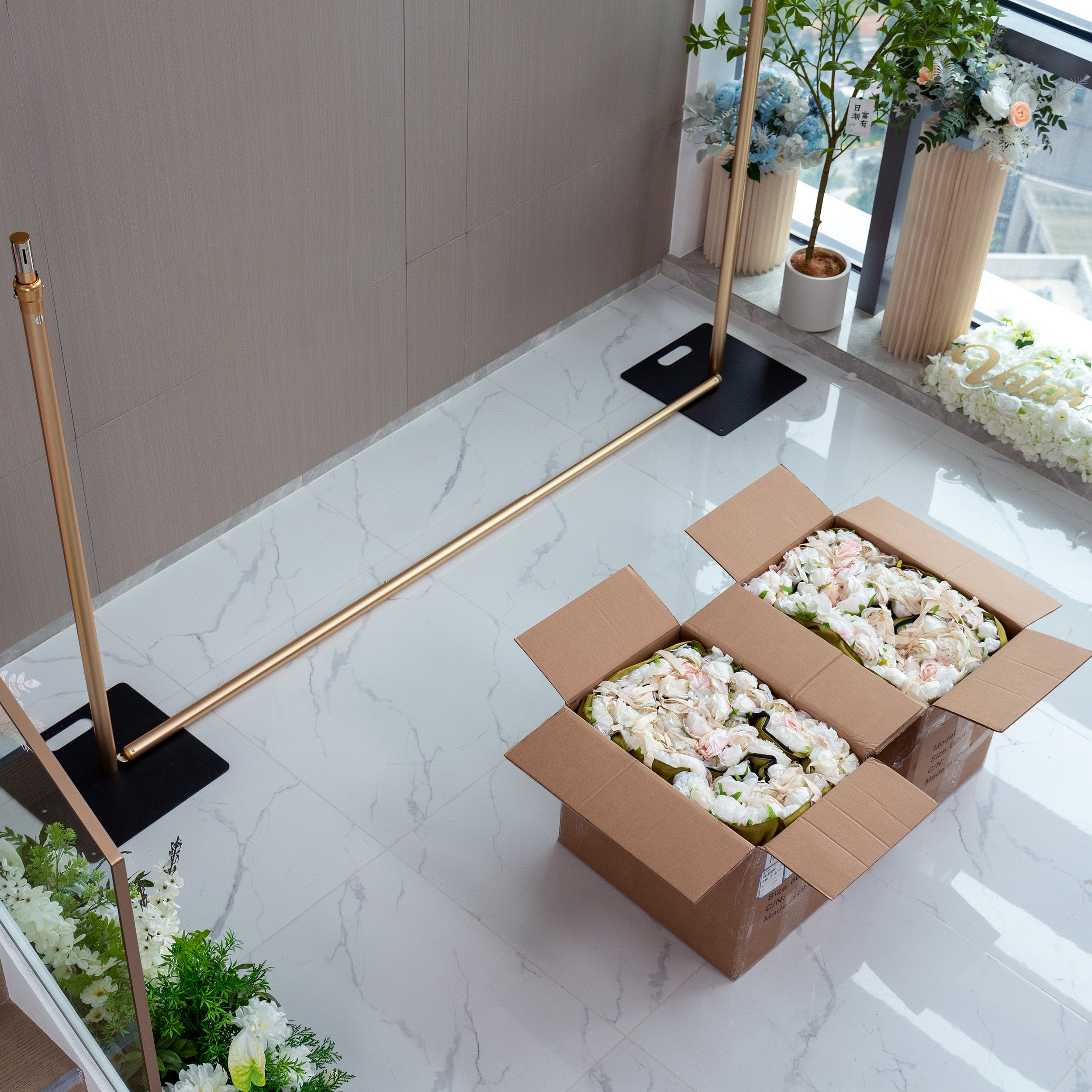 This image shows the setup process of a floral wall. Two golden poles are positioned on a marble floor, with two cardboard boxes filled with white and light - pink flowers placed between them. The boxes are open, revealing the flowers inside, which are likely being prepared for arrangement on the wall. The scene also includes some potted plants and additional floral decorations in the background, suggesting a floral - themed event setup.
