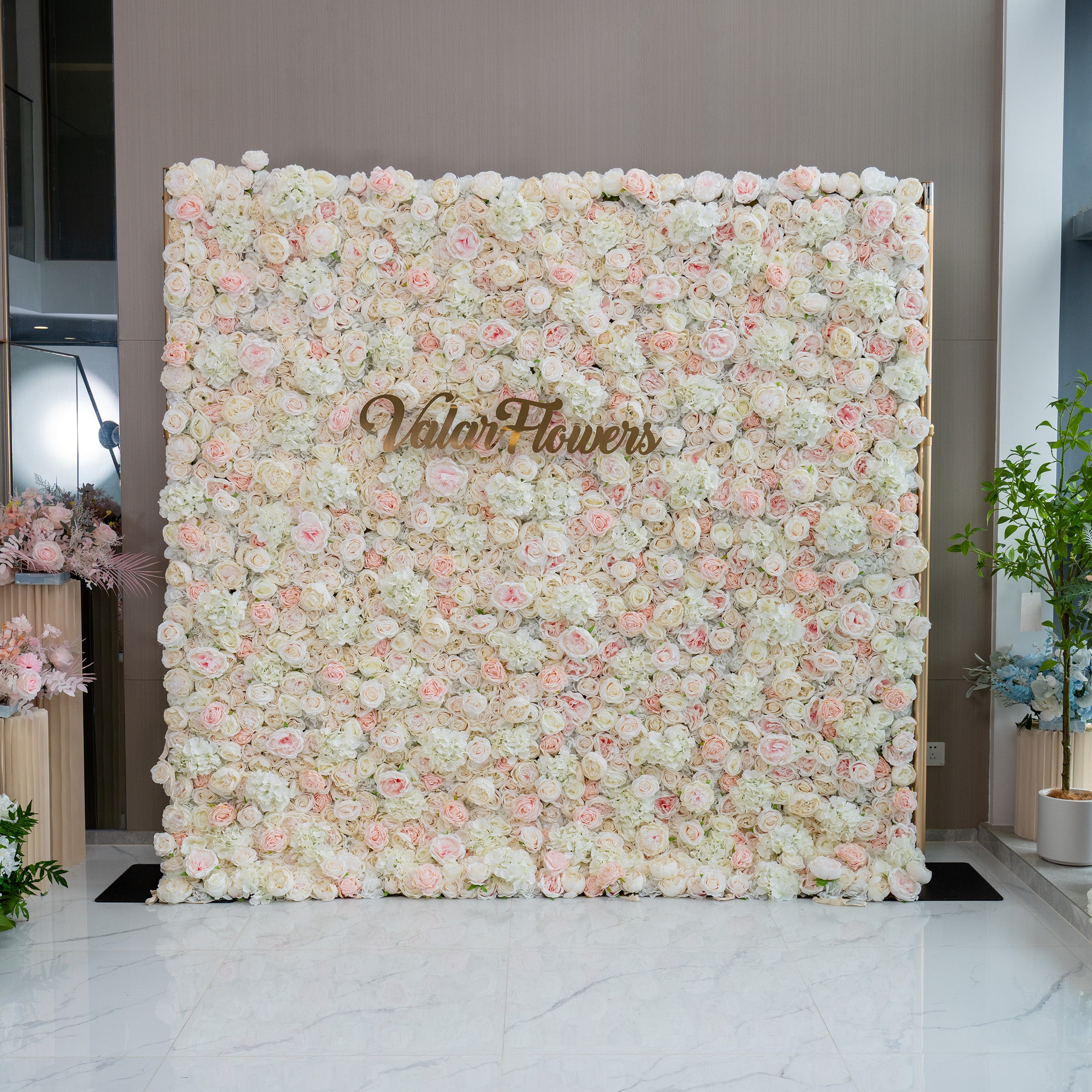 This image showcases a floral wall adorned with a dense arrangement of light pink and white flowers. The wall features the brand name "ValarFlowers" prominently displayed in the center. The overall ambiance is soft, romantic, and inviting, making it ideal for events such as weddings, bridal showers, or romantic celebrations.