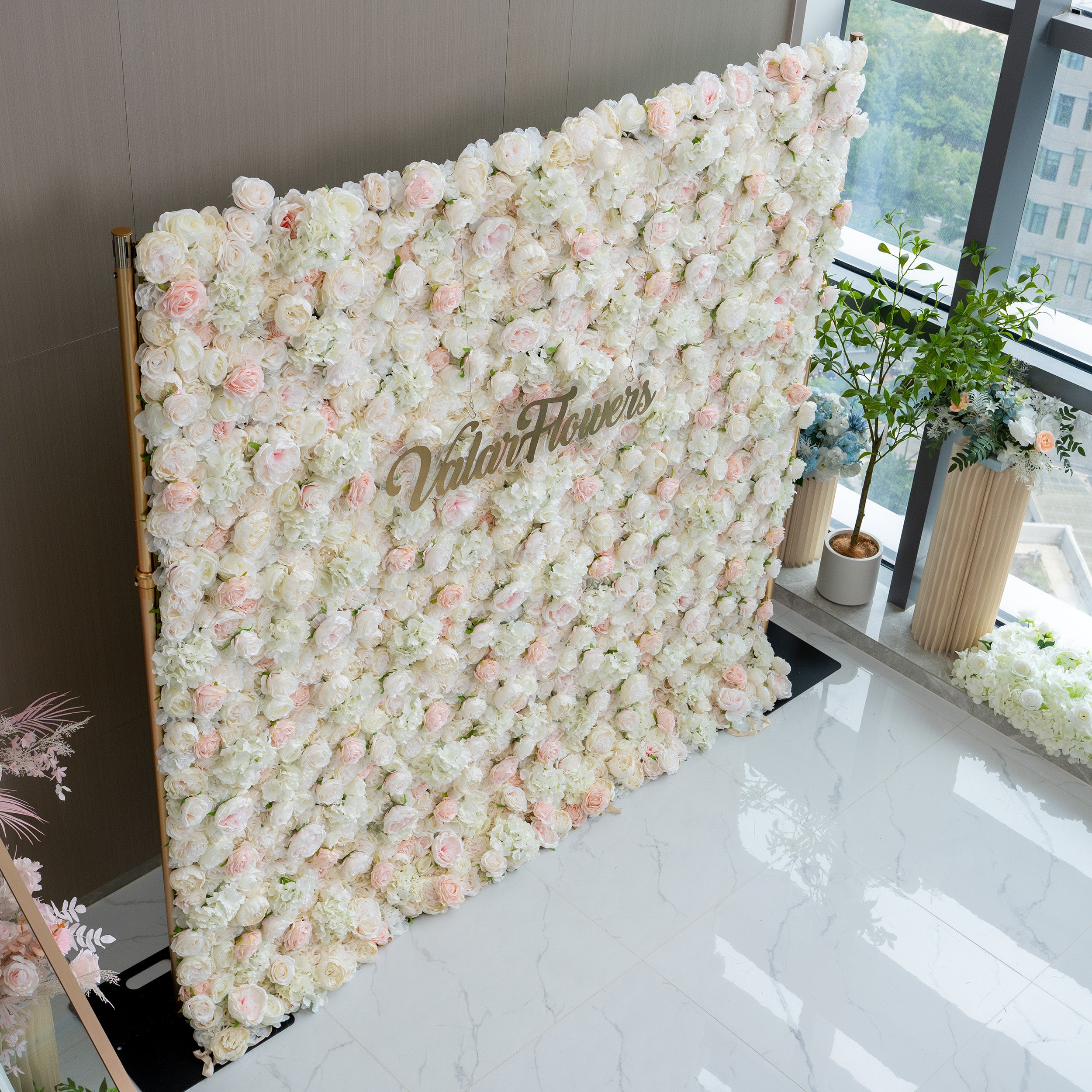 This image showcases a floral wall adorned with a dense arrangement of light pink and white flowers. The wall prominently displays the text "ValarFlowers" in the center. The overall ambiance is soft, romantic, and inviting, making it ideal for events such as weddings, bridal showers, or romantic celebrations.