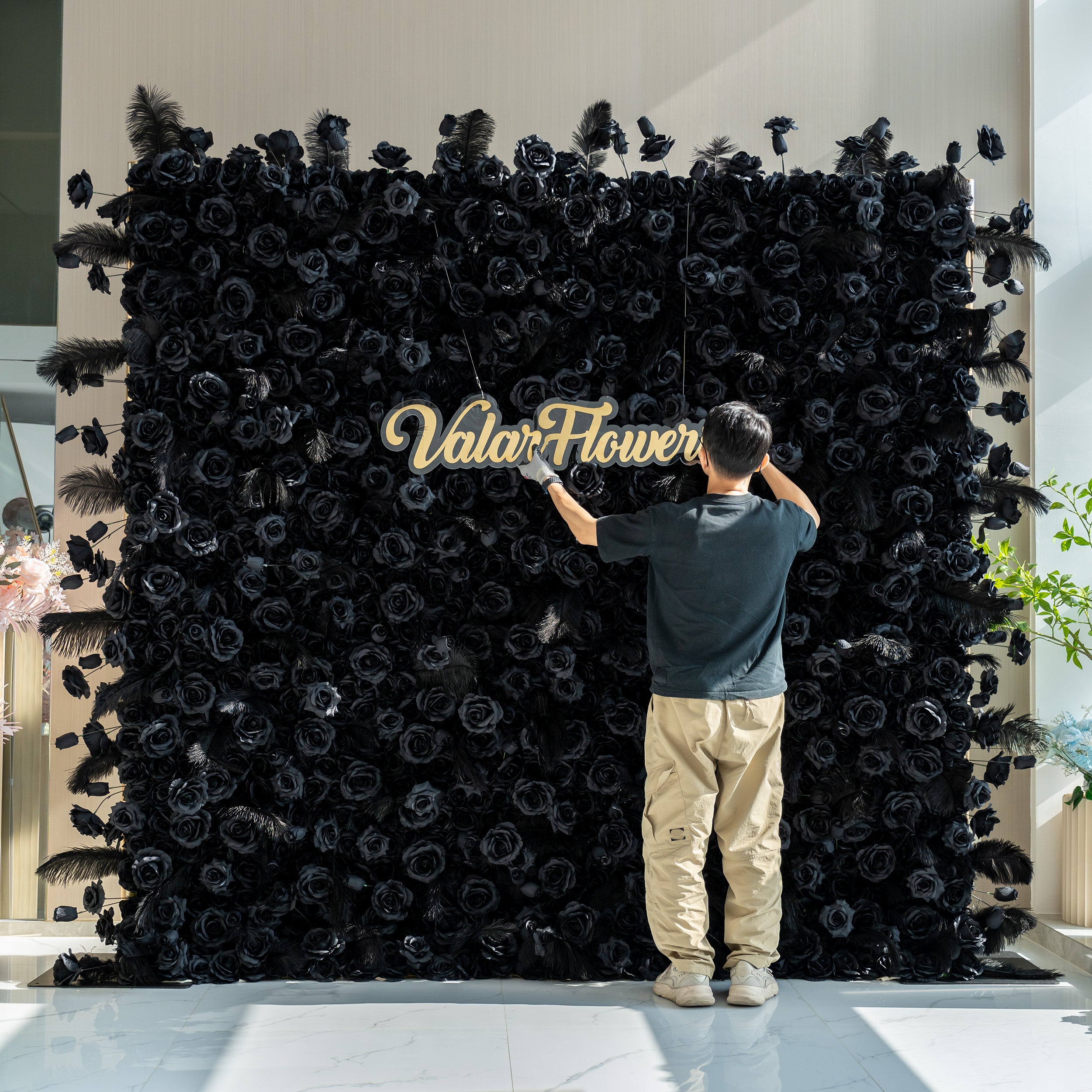 A person is setting up a sign for "ValarFlowers" in front of a stunning black rose wall. The wall, adorned with feathers, creates a dramatic and elegant display.