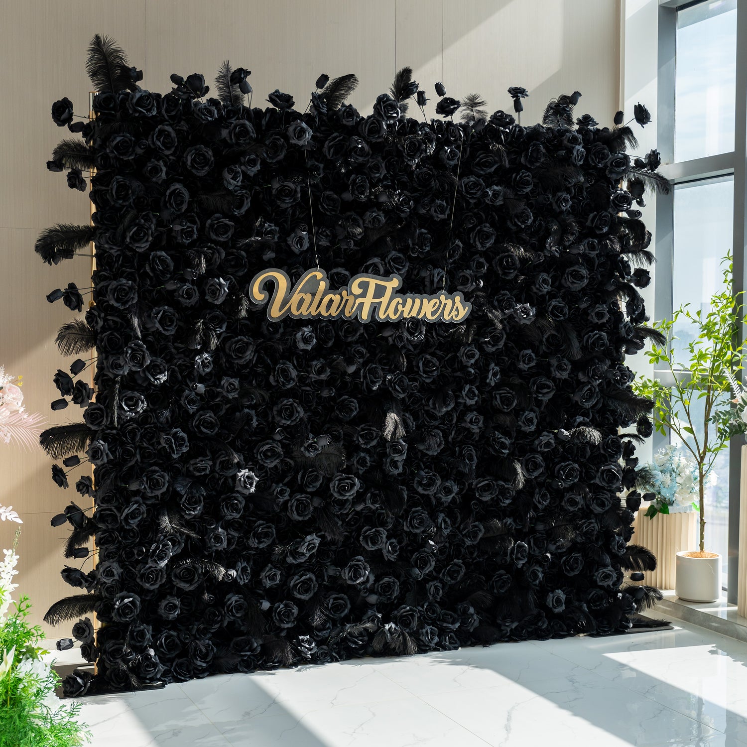 The black rose wall with the "ValarFlowers" sign creates a dramatic and luxurious atmosphere. The contrast of the dark roses and the light background, along with the feather accents, adds an air of elegance and sophistication.