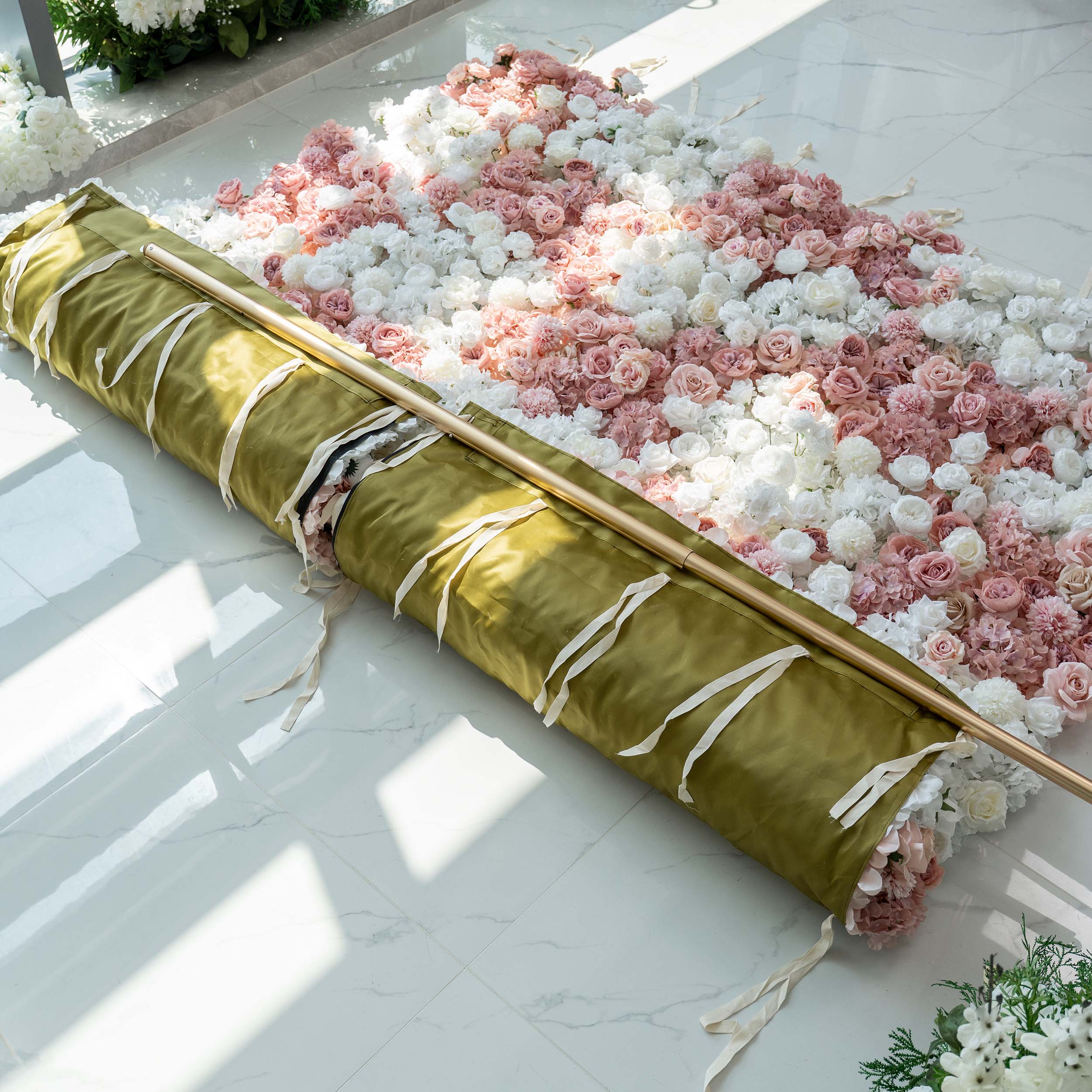 An 8x8ft rolled - up floral wall on a marble floor, featuring a mix of white and pink artificial roses and hydrangeas, with a green fabric backing and white ties.