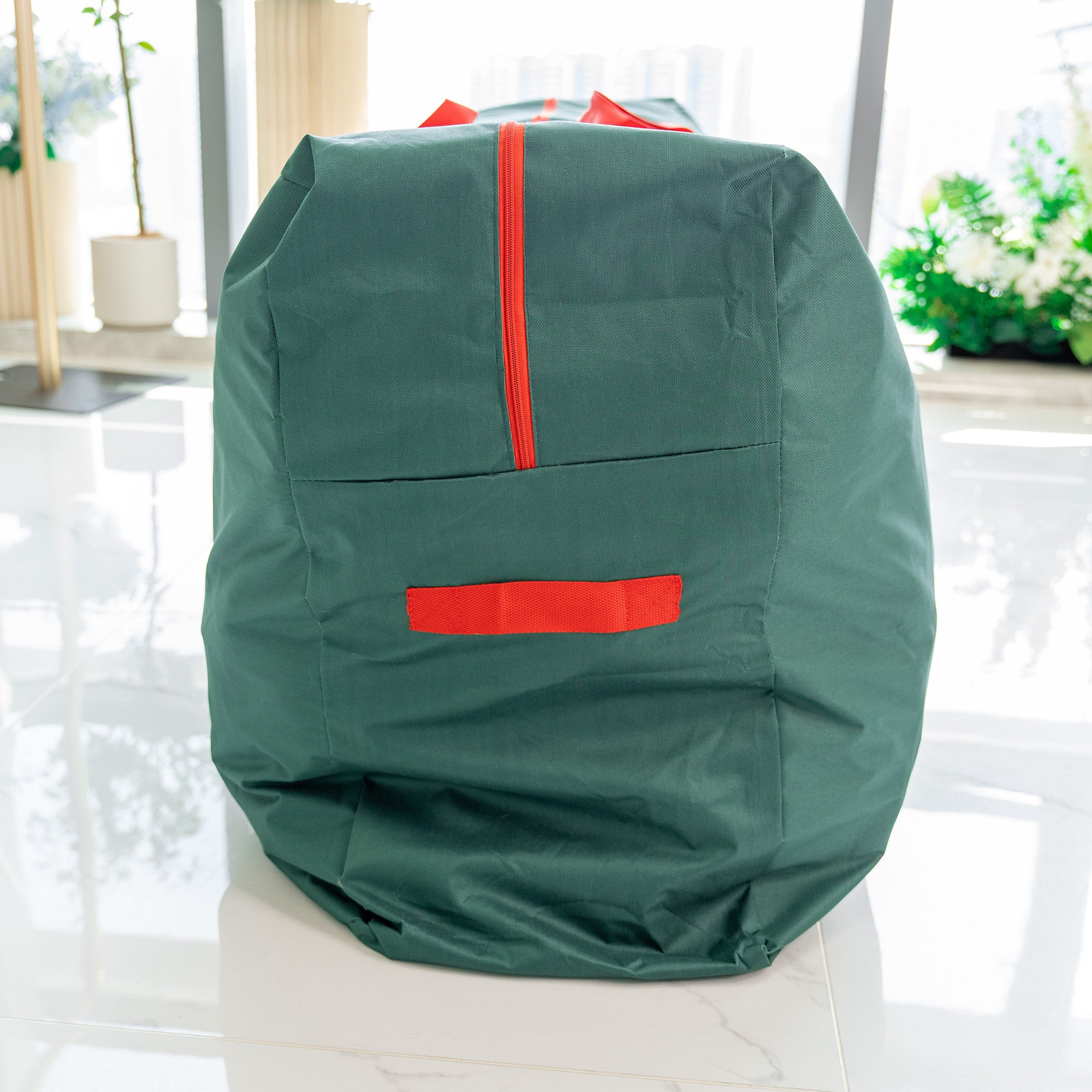 This image shows the side view of a storage bag for flower walls. The bag is in a dark green color. There is a prominent red zipper running vertically along the side, which is likely used for opening and closing the bag. A red handle is attached to the side, providing a means for carrying. The background includes a bright indoor setting with some greenery and potted plants, suggesting a clean and well - lit environment.