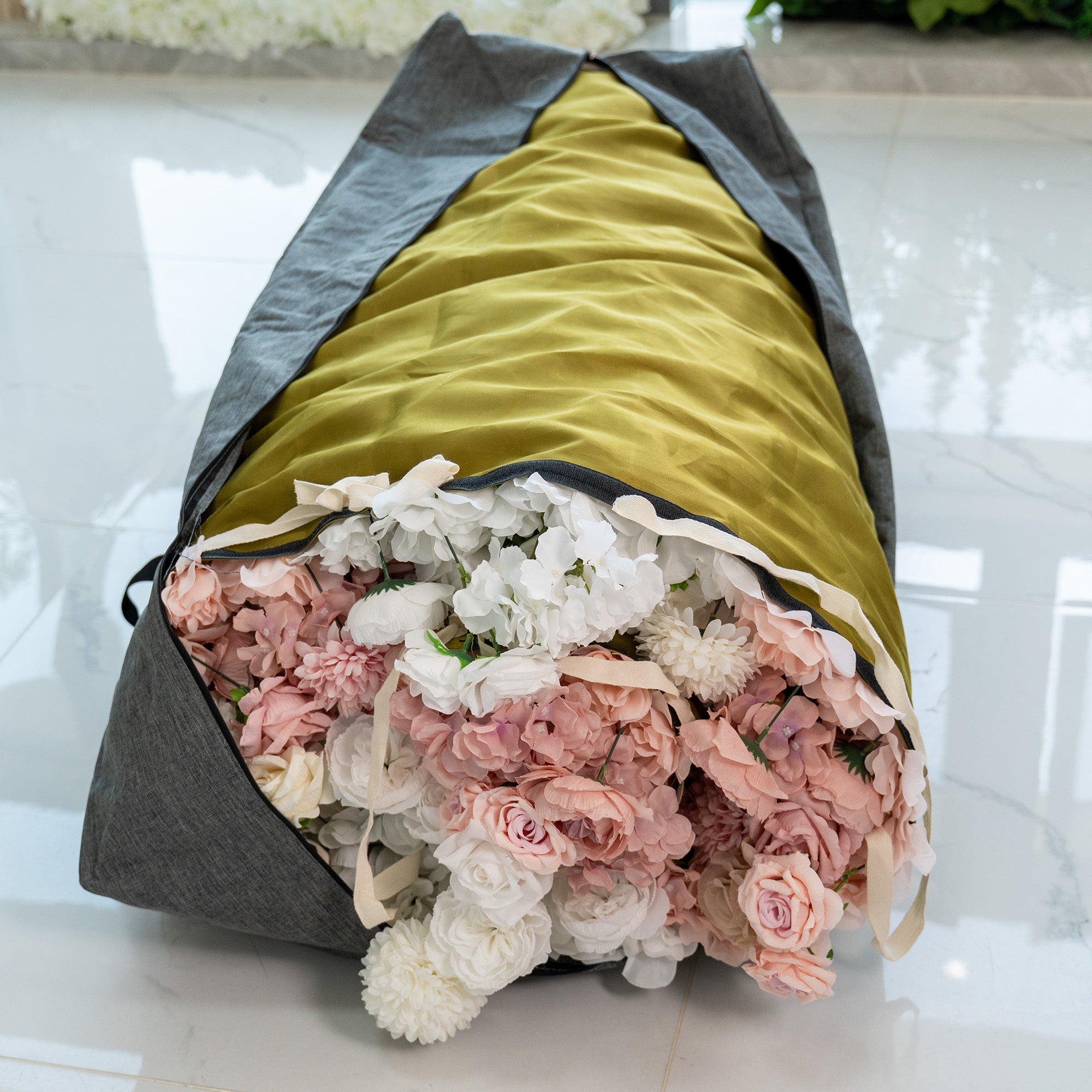 This image shows a storage bag designed for flower walls. The bag has a gray exterior with a mustard - colored interior. It is partially open, revealing a generous collection of artificial flowers in soft hues of pink, white, and light purple. The flowers appear to be well - packed inside. The bag also has straps, indicating it is portable, making it convenient for storing and transporting flower wall decorations.