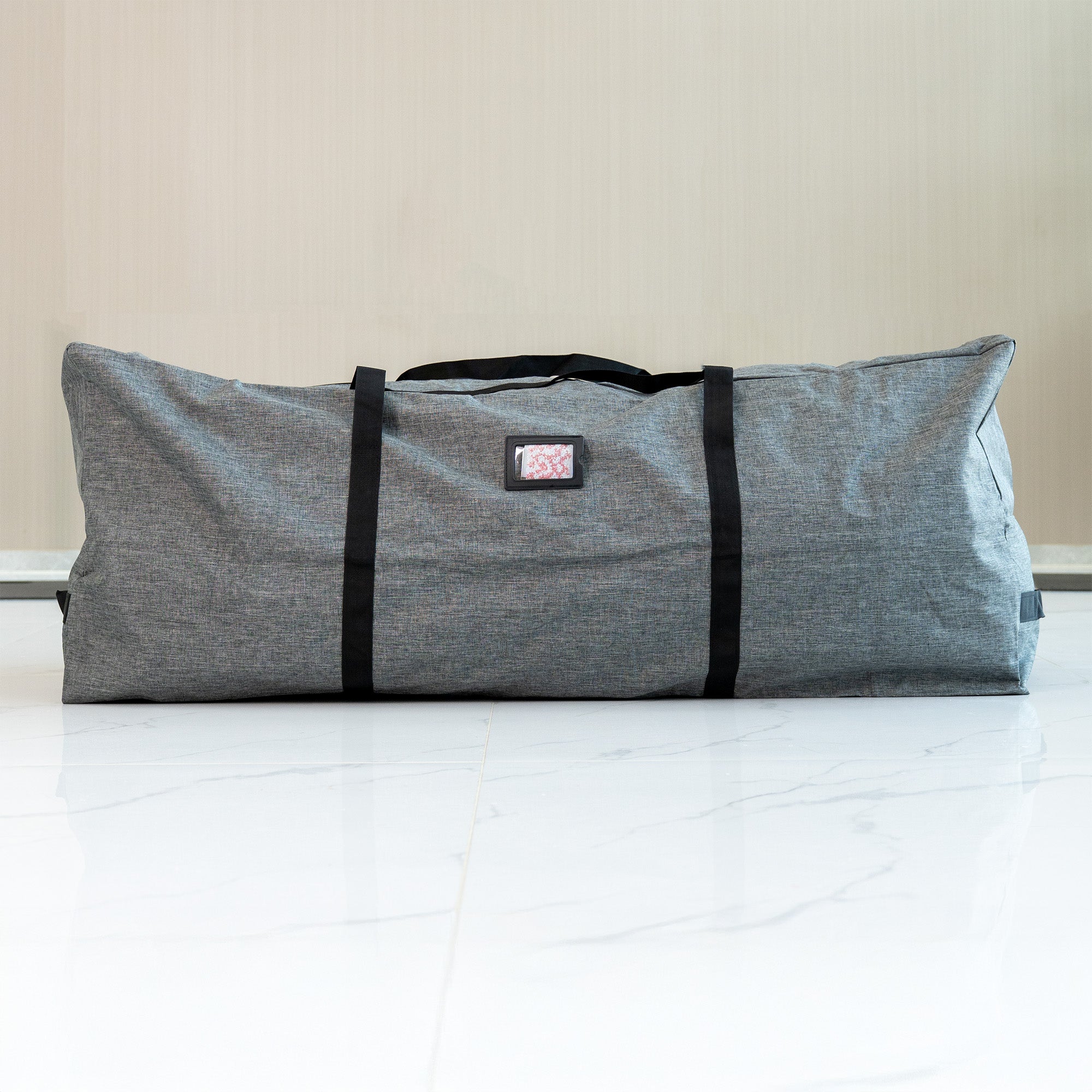This is a storage bag for flower walls. It is large and rectangular, with a heathered gray fabric exterior. The bag is equipped with sturdy black carrying straps and features a small rectangular label patch with a pink - patterned insert. It is placed on a white marble - tiled floor against a neutral - colored wall, suggesting a clean and modern setting. The bag is designed for convenient storage and transport of flower wall decorations.