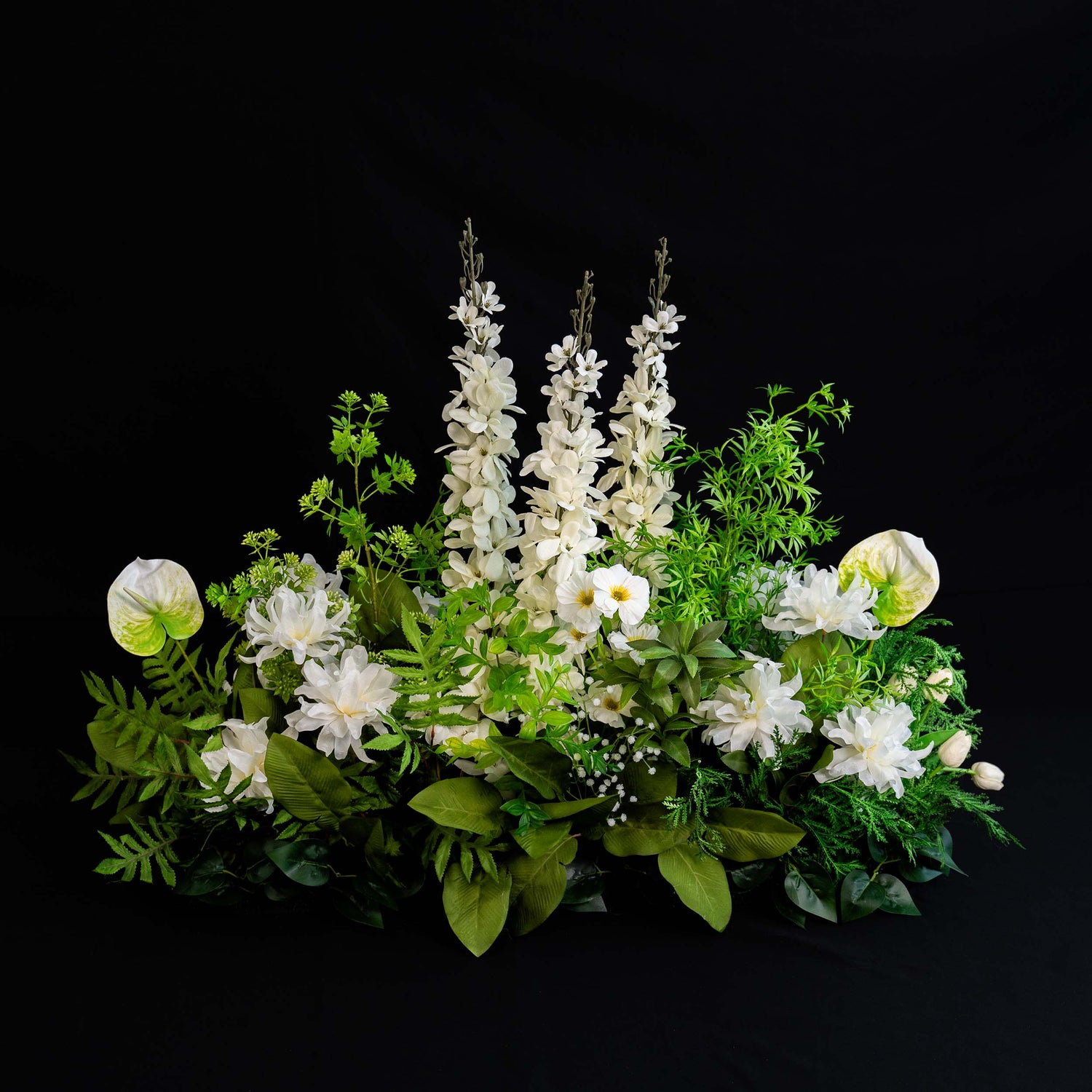A floral arrangement features tall white blooms amidst an array of white flowers and lush green foliage, set against a stark black background for a striking contrast.