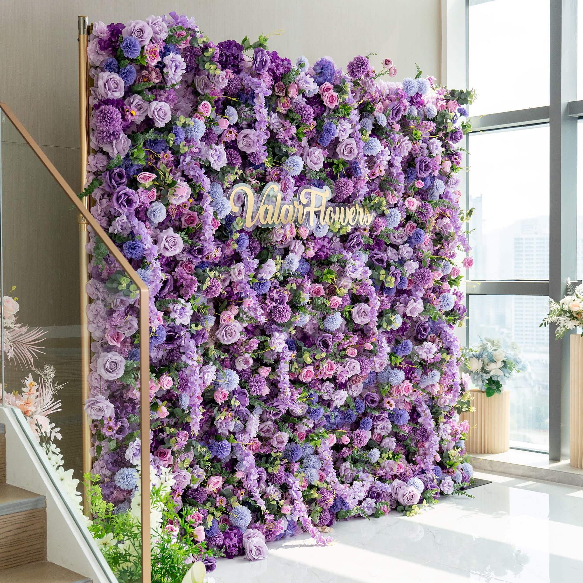 This image features a 2.4 x 2.4m floral wall supported by a golden stand. The wall is densely packed with a variety of artificial flowers in rich shades of purple, ranging from deep violet to light lavender, along with hints of pink and green. Prominent among the blooms are roses, hydrangeas, and chrysanthemum - like flowers. The "ValarFlowers" logo is displayed in the center, adding a branding element to this opulent and romantic floral backdrop.