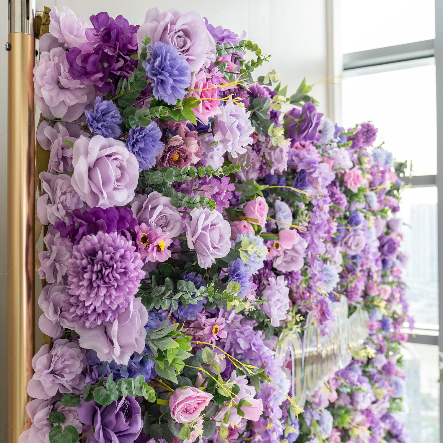 This image showcases a lush floral arrangement on a wall. It features an array of artificial flowers in various shades of purple, from deep violet to light lavender, along with hints of pink. Prominent flowers include roses, large - petaled blooms resembling peonies or chrysanthemums, and clusters of smaller flowers. Interspersed greenery adds a fresh touch, creating a rich, romantic, and visually appealing display.