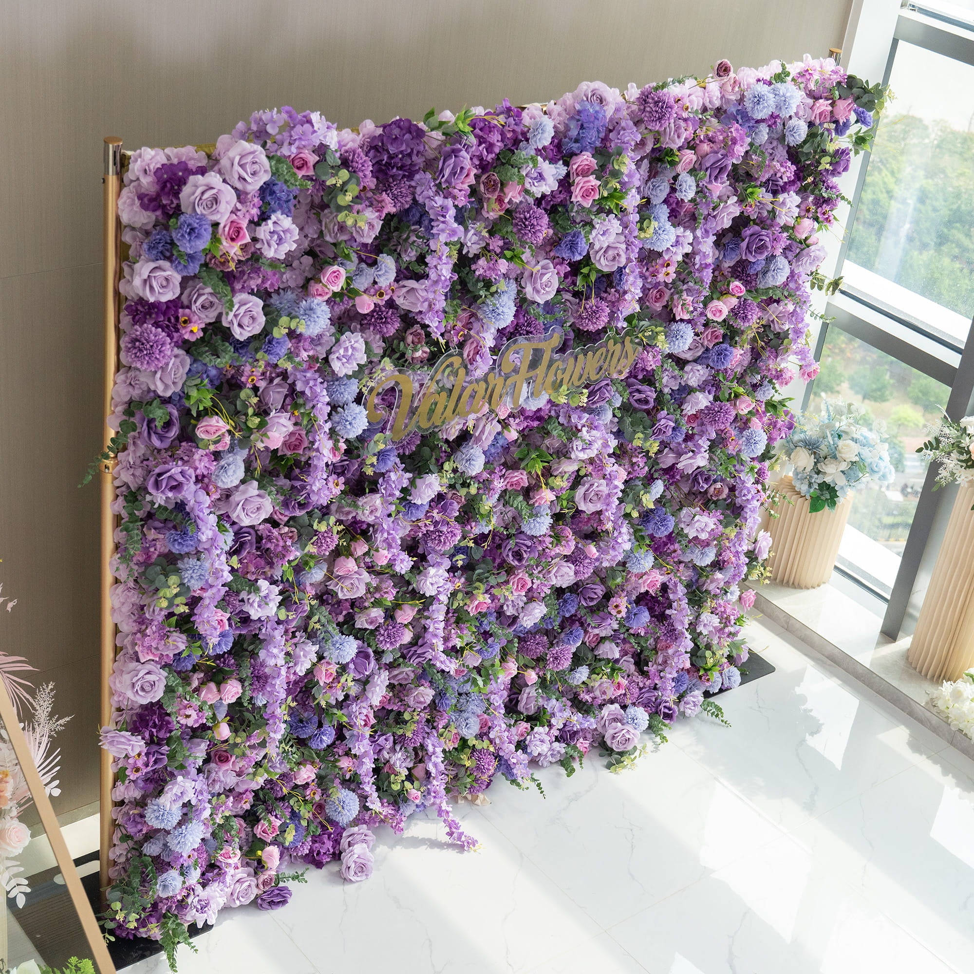 This image showcases a lush, vibrant floral wall supported by a golden frame. The wall is densely adorned with an array of artificial flowers in various shades of purple, from deep violet to light lavender, along with hints of pink and green. Prominent flowers include roses, hydrangeas, and chrysanthemum - like blooms. In the center, the text "ValarFlowers" in a cursive, golden font adds a touch of elegance, making it perfect for events or photo backdrops.