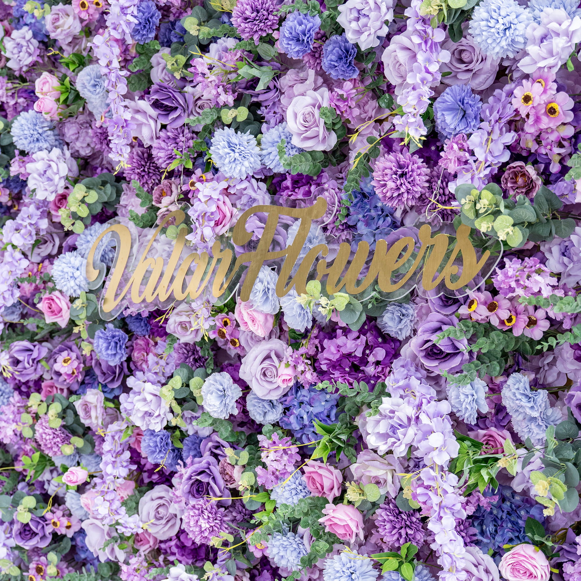 This image showcases a rich, textured floral wall with a captivating blend of artificial flowers in various shades of purple, from deep violet to light lavender, along with hints of pink and blue. Prominent flowers include roses, hydrangeas, and chrysanthemum - like blooms, interspersed with greenery. In the center, the golden text "ValarFlowers" adds an elegant touch, enhancing the overall romantic and opulent aesthetic.