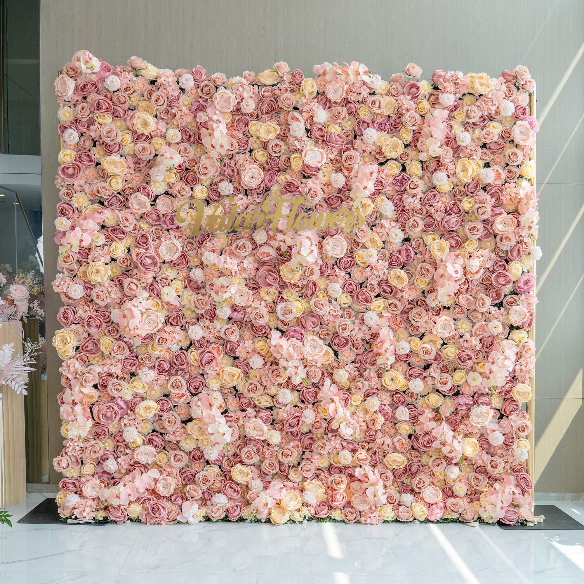 The image features a large flower wall backdrop filled with artificial roses in shades of pink, blush, and cream. In the center, the branding “Valarflowers” in gold - toned letters stands out. The style is opulent and romantic.