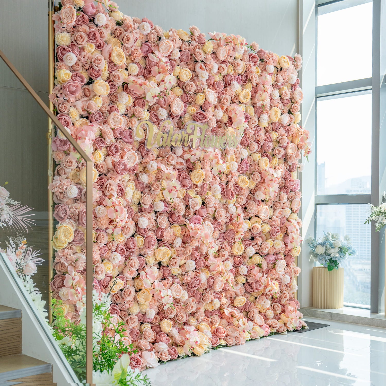 The image displays a large flower wall densely covered in artificial roses in soft pink, blush, and cream tones. The “Valarflowers” branding in gold is centered. It has a luxurious, romantic style, set indoors by a staircase.
