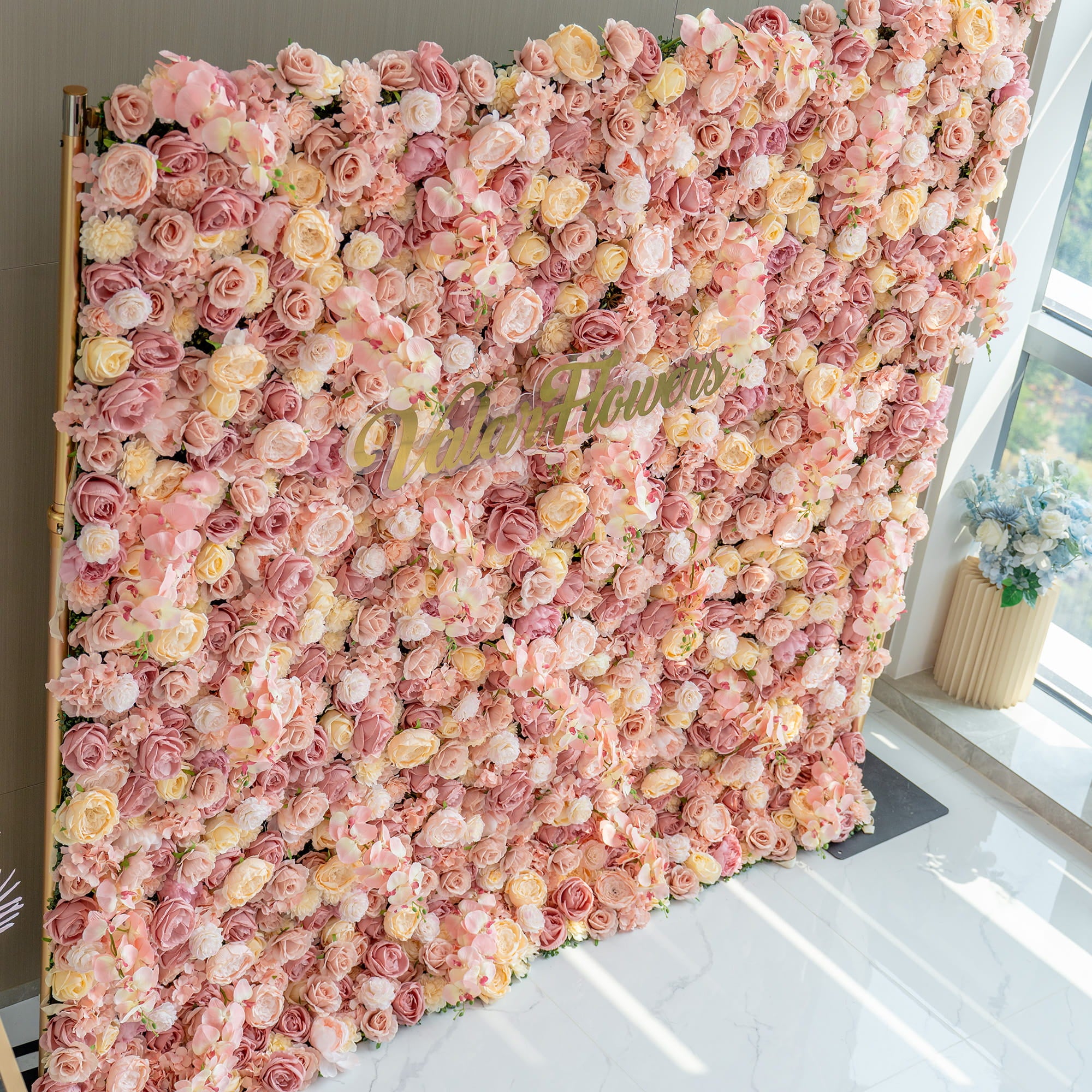 This image showcases a large flower wall filled with artificial roses in soft pink, blush, and cream tones. The "Valarflowers" branding in gold stands out in the center. The style is luxurious and romantic.