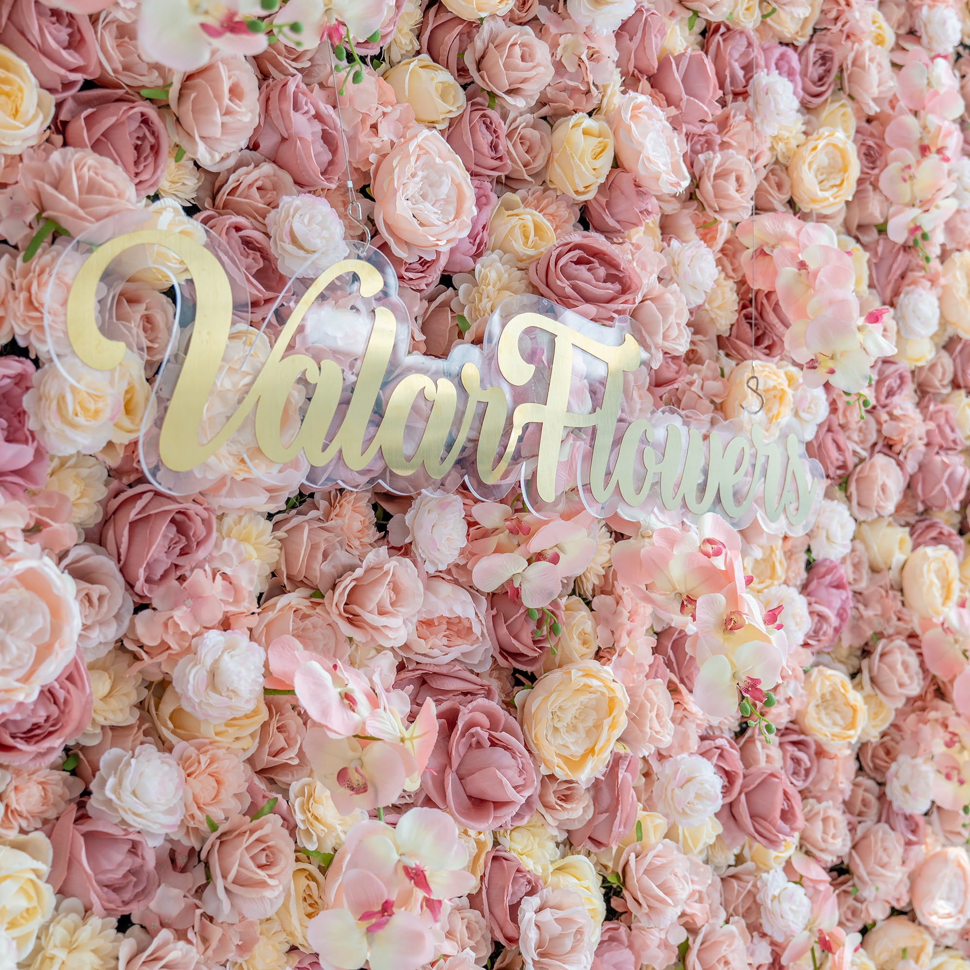 The image shows a flower wall densely packed with artificial roses and orchids in various pastel shades like pink, peach, and cream. The branding “ValarFlowers” in gold on a clear base stands out. The style is sumptuous and romantic.