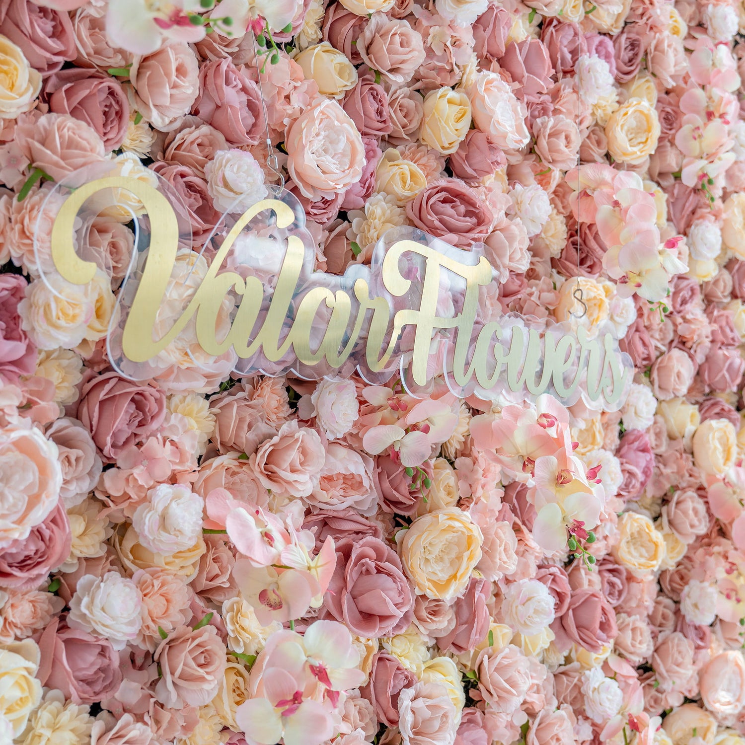 The image shows a flower wall densely packed with artificial roses and orchids in various pastel shades like pink, peach, and cream. The branding “ValarFlowers” in gold on a clear base stands out. The style is sumptuous and romantic.