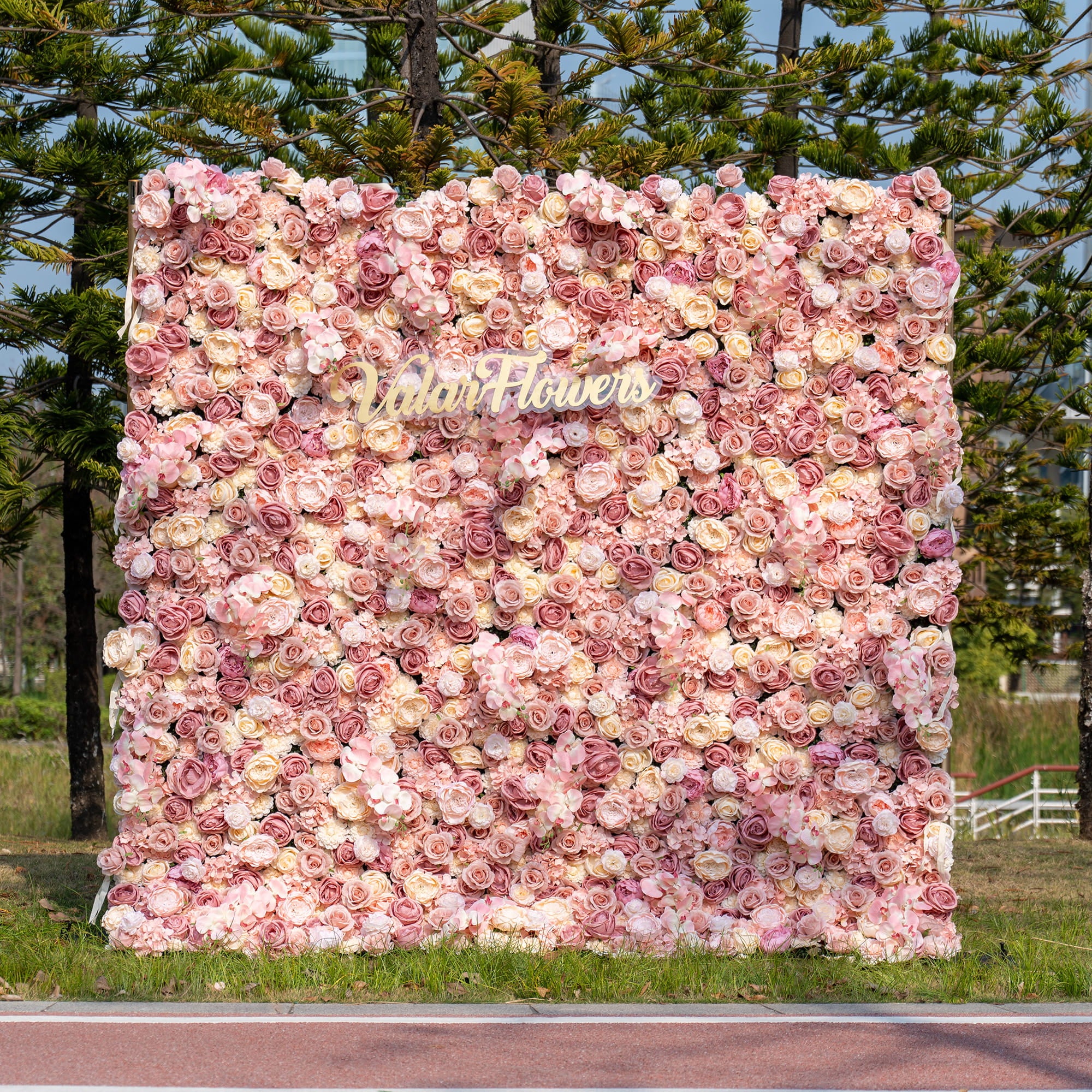 The image shows an outdoor flower wall mainly made of artificial roses in soft pink, peach, and cream colors. The “ValarFlowers” logo is in the middle. It has a romantic and elegant style.