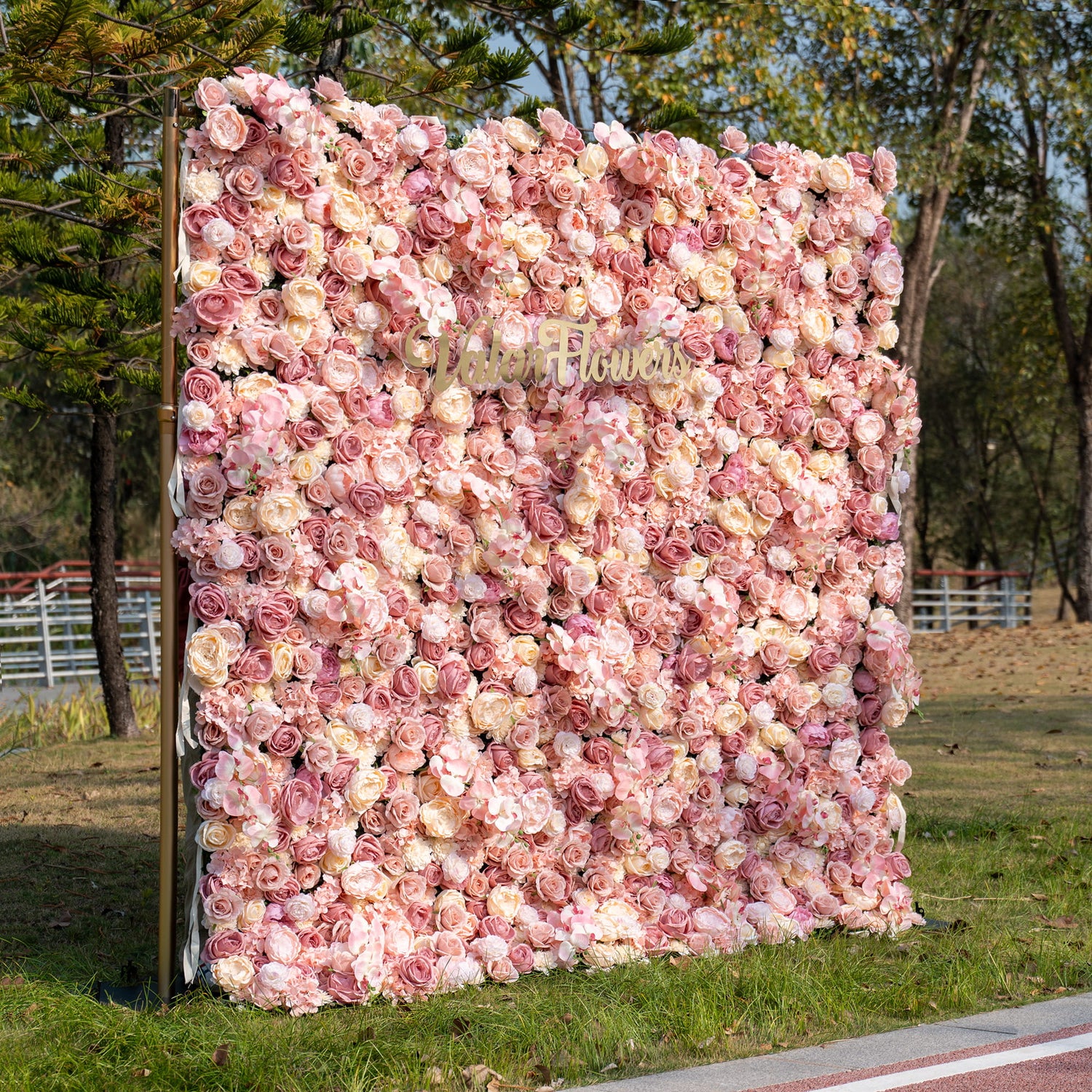 This outdoor flower wall is densely covered with artificial roses in soft pink, peach, and cream hues. The “ValarFlowers” logo is prominently displayed in the center, creating a romantic and elegant style.