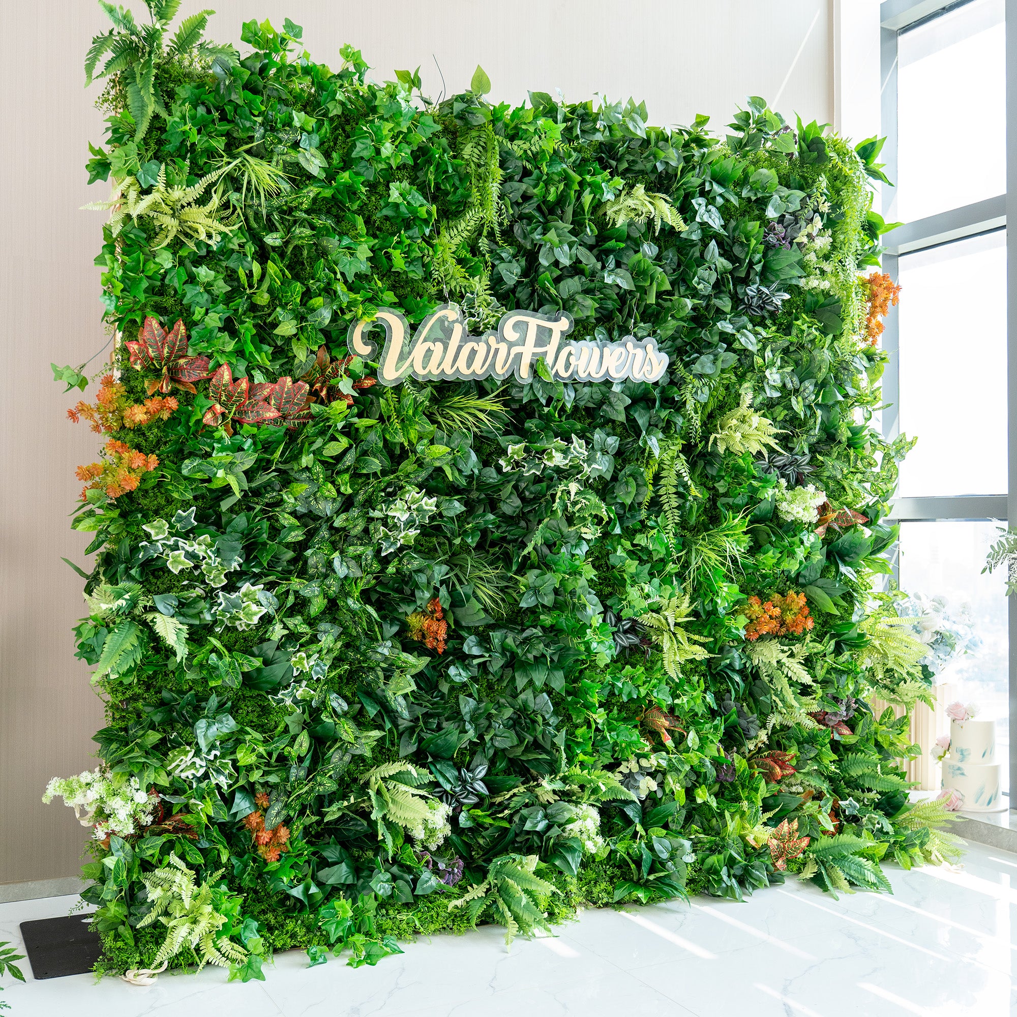 This image showcases a lush, vibrant green wall created by ValarFlowers. The wall is densely packed with a variety of artificial plants, including different shades of green foliage such as ivy, ferns, and broad - leafed plants. There are also splashes of color from orange and reddish - brown plants, adding a lively contrast. The overall effect is a rich, natural - looking greenery wall, perfect for event backdrops or decorative purposes.