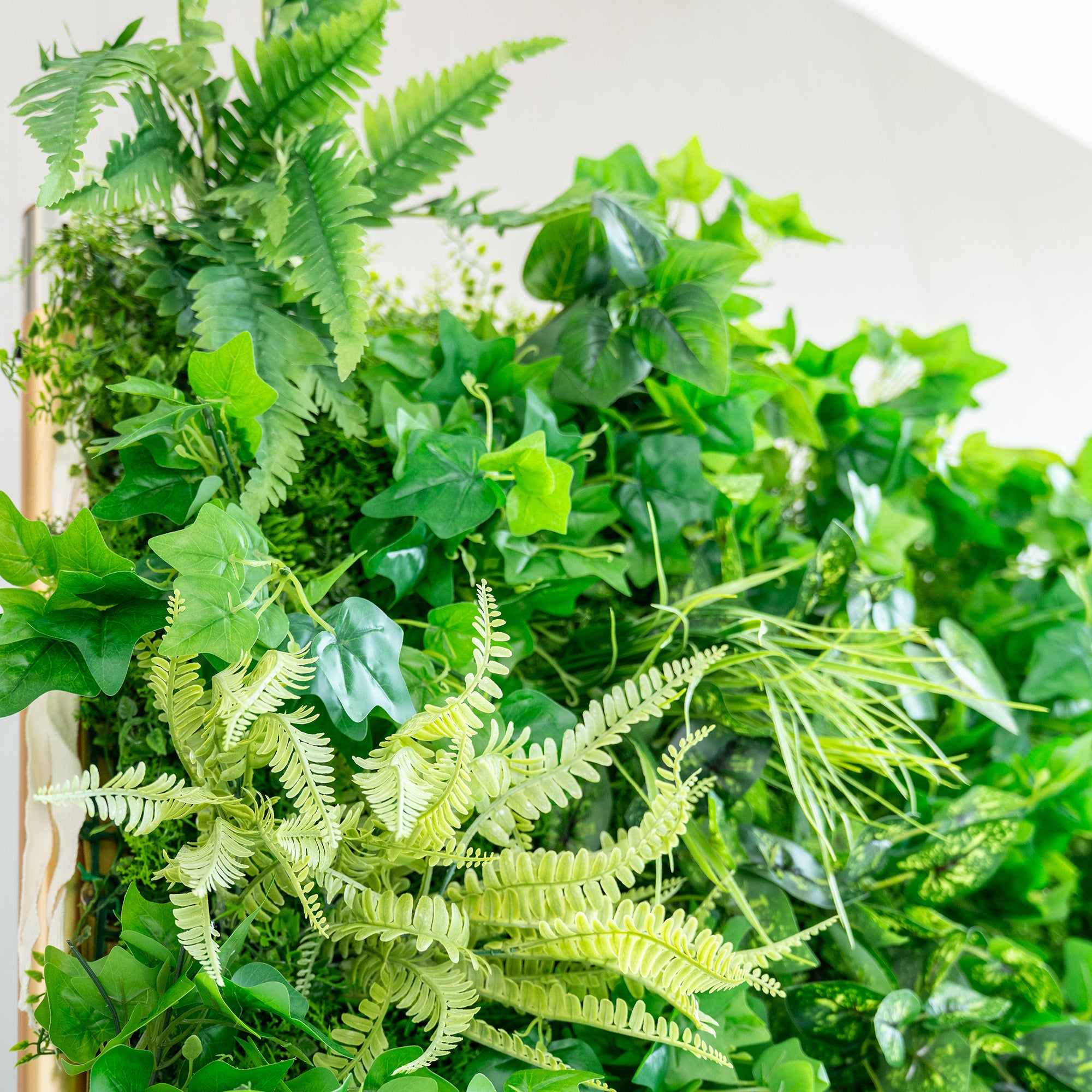 This image showcases a lush arrangement of artificial greenery. It prominently features various types of ferns in different shades of green, from light lime to deeper emerald hues. Interspersed among the ferns are broad - leafed plants, adding texture and variety to the display. The overall effect is a vibrant, natural - looking cluster of foliage, ideal for decorative purposes, such as event backdrops or interior decor.