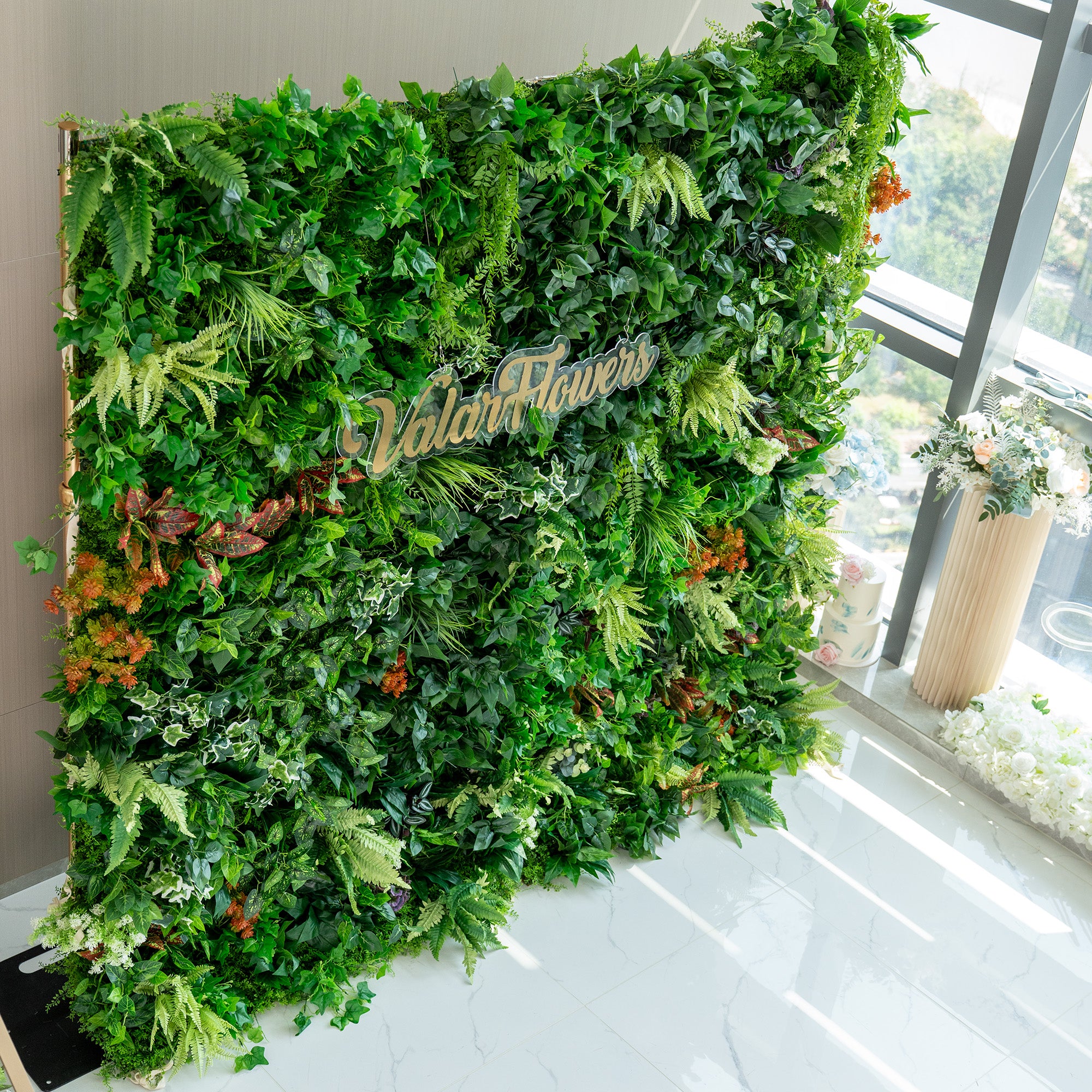 This image features a lush, artificial green wall by ValarFlowers. The wall is densely packed with a variety of faux plants, including different shades of green foliage such as ivy - like leaves, ferns, and broad - leafed plants. There are also splashes of color from orange and red - tinged plants, adding a vibrant contrast. The backdrop is set indoors near a window, with some floral arrangements visible nearby, enhancing the fresh and natural ambiance.