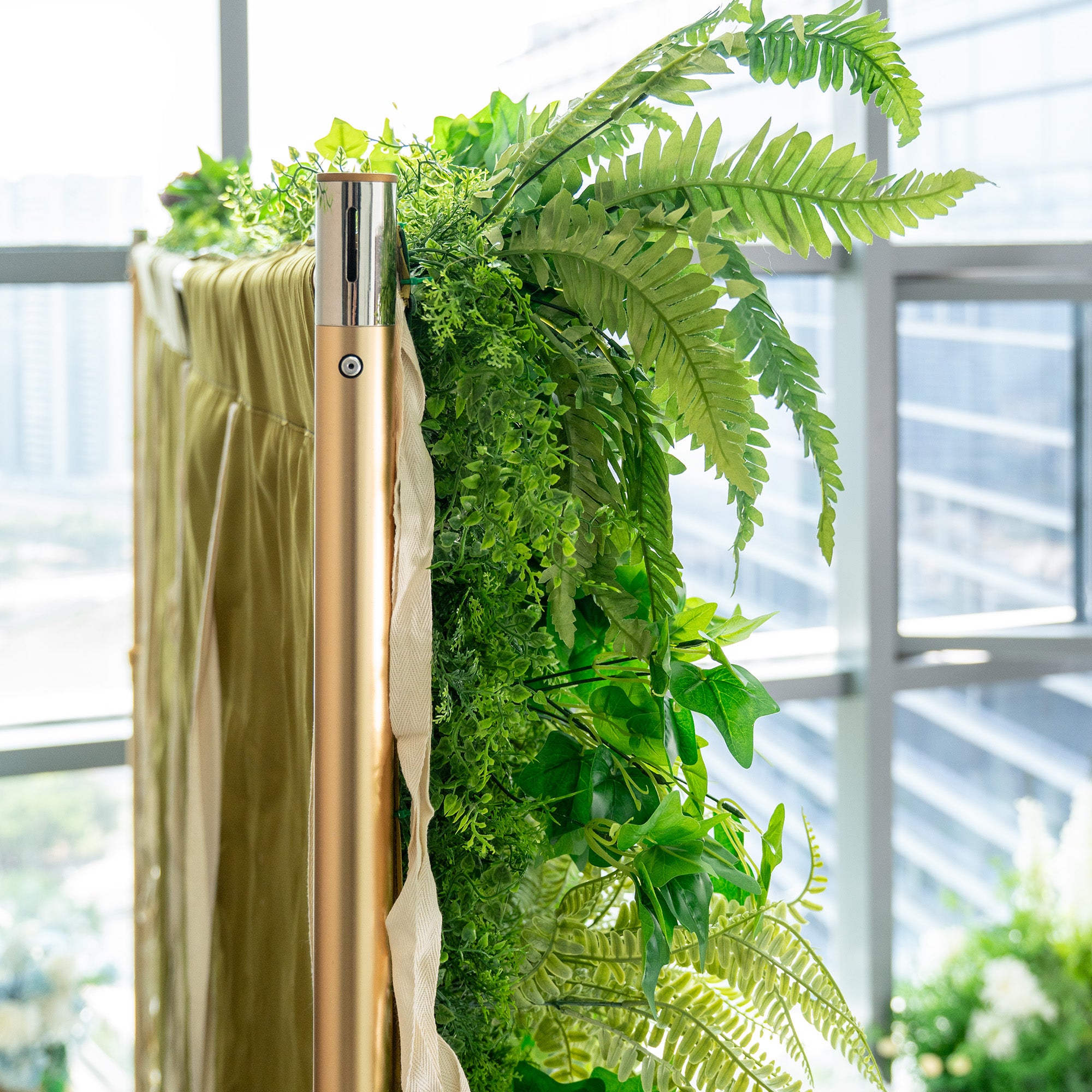This image captures a close - up view of a lush greenery arrangement attached to a structure. Prominently featured are vibrant green fern leaves, adding a touch of nature. A metallic golden pole with a sleek design is visible, likely part of the support structure. Behind the greenery, a mustard - colored fabric is partially seen, suggesting it might be part of a backdrop or enclosure. The background shows a blurred view of a modern building through large windows, indicating an indoor setting with natural li